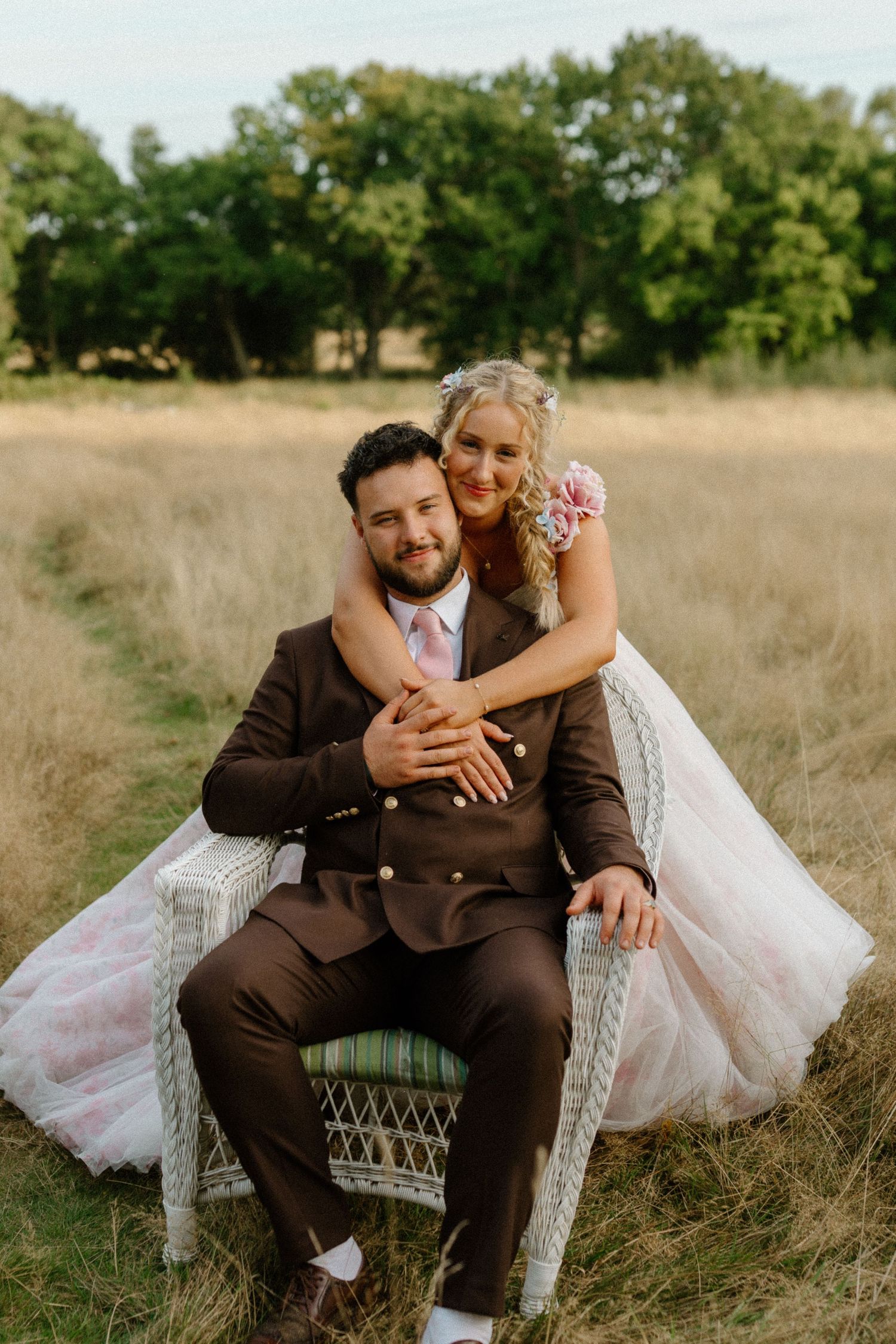 A smiling bride leans over her groom seated in a wicker chair in a sun-drenched countryside field.