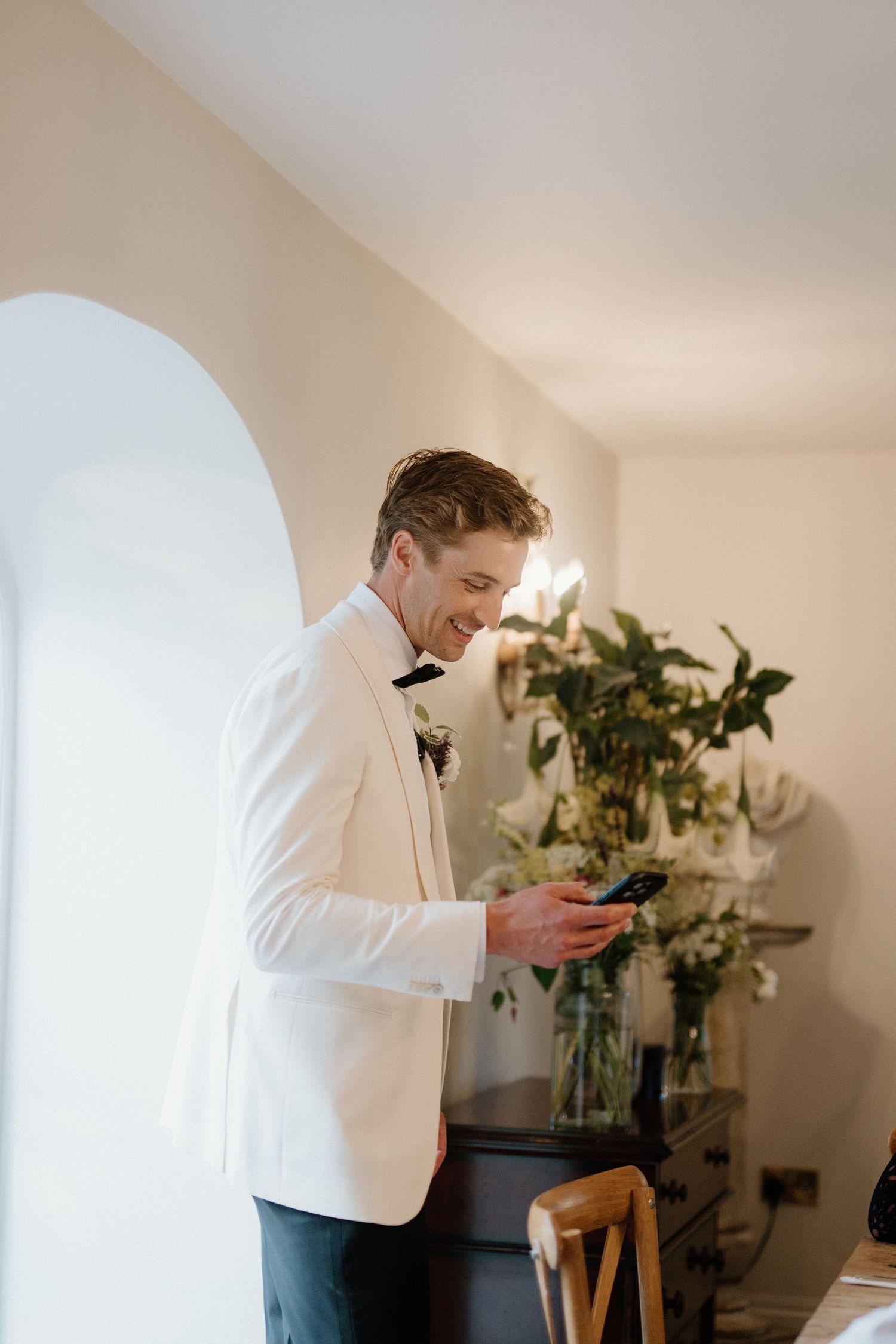 Smiling groom in white tuxedo jacket checking phone before wedding ceremony.