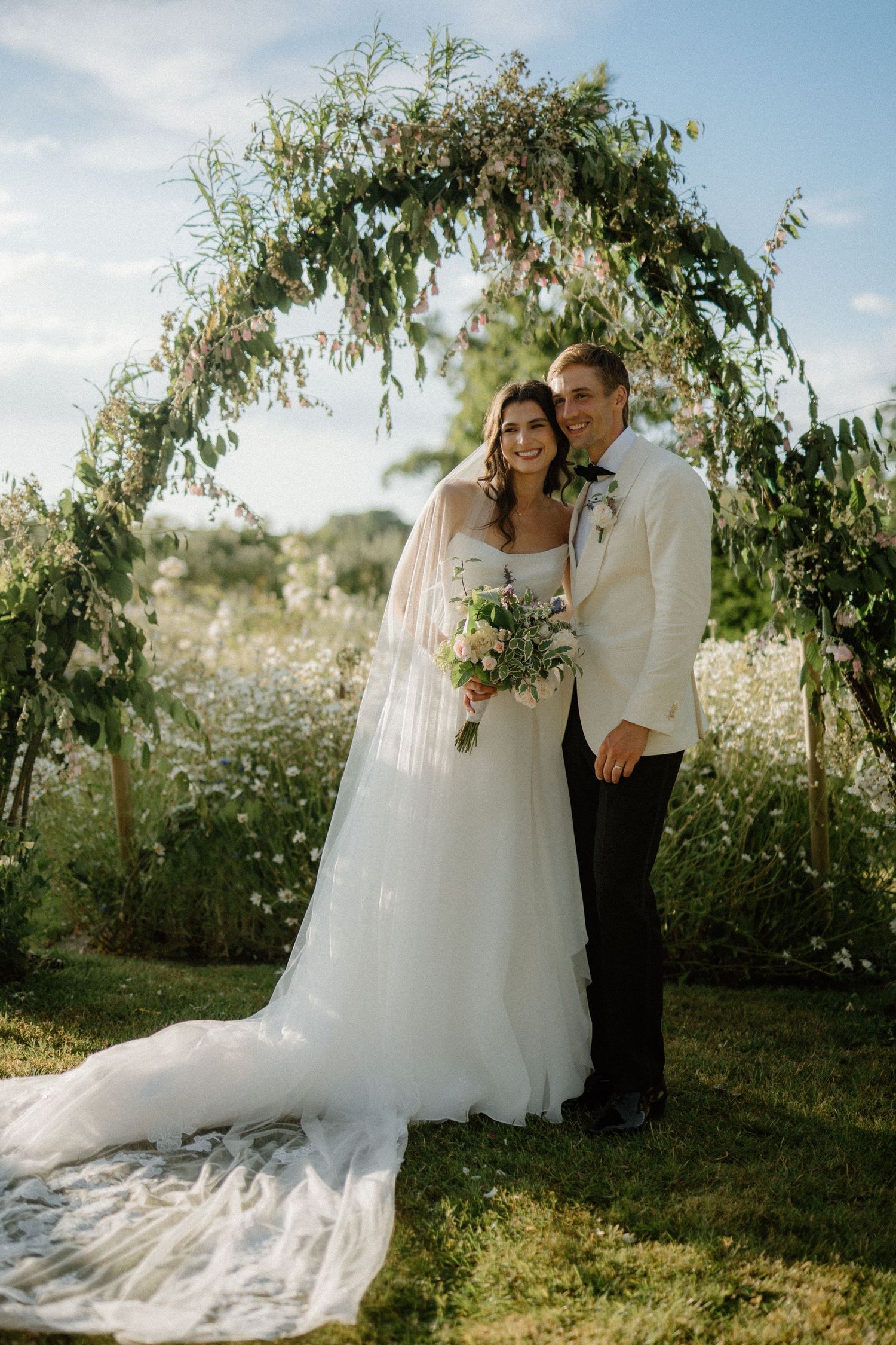 Bride and groom posing together under greenery arch in wildflower garden after outdoor ceremony.