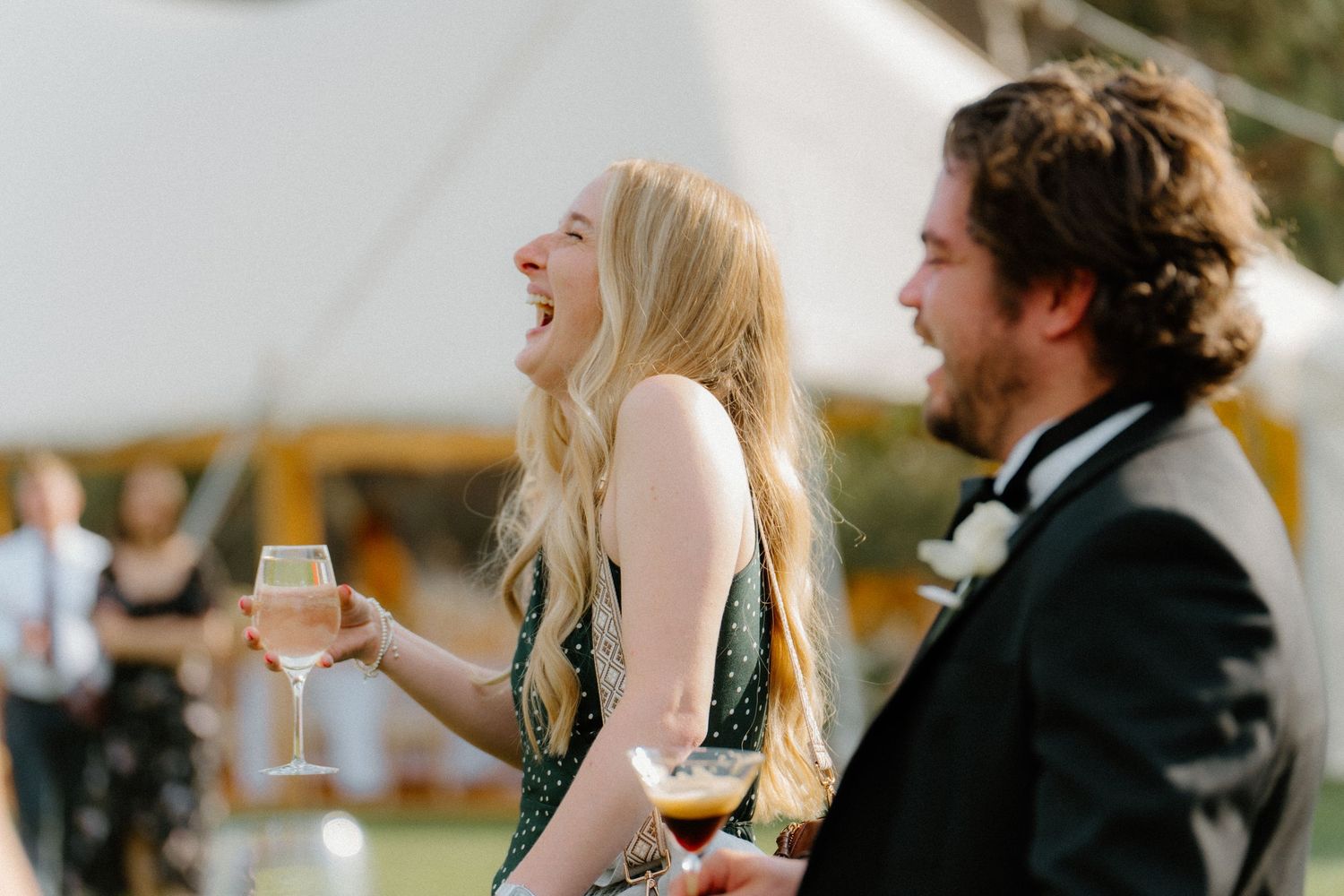 Wedding guest in green dress laughing while holding champagne glass at outdoor reception.