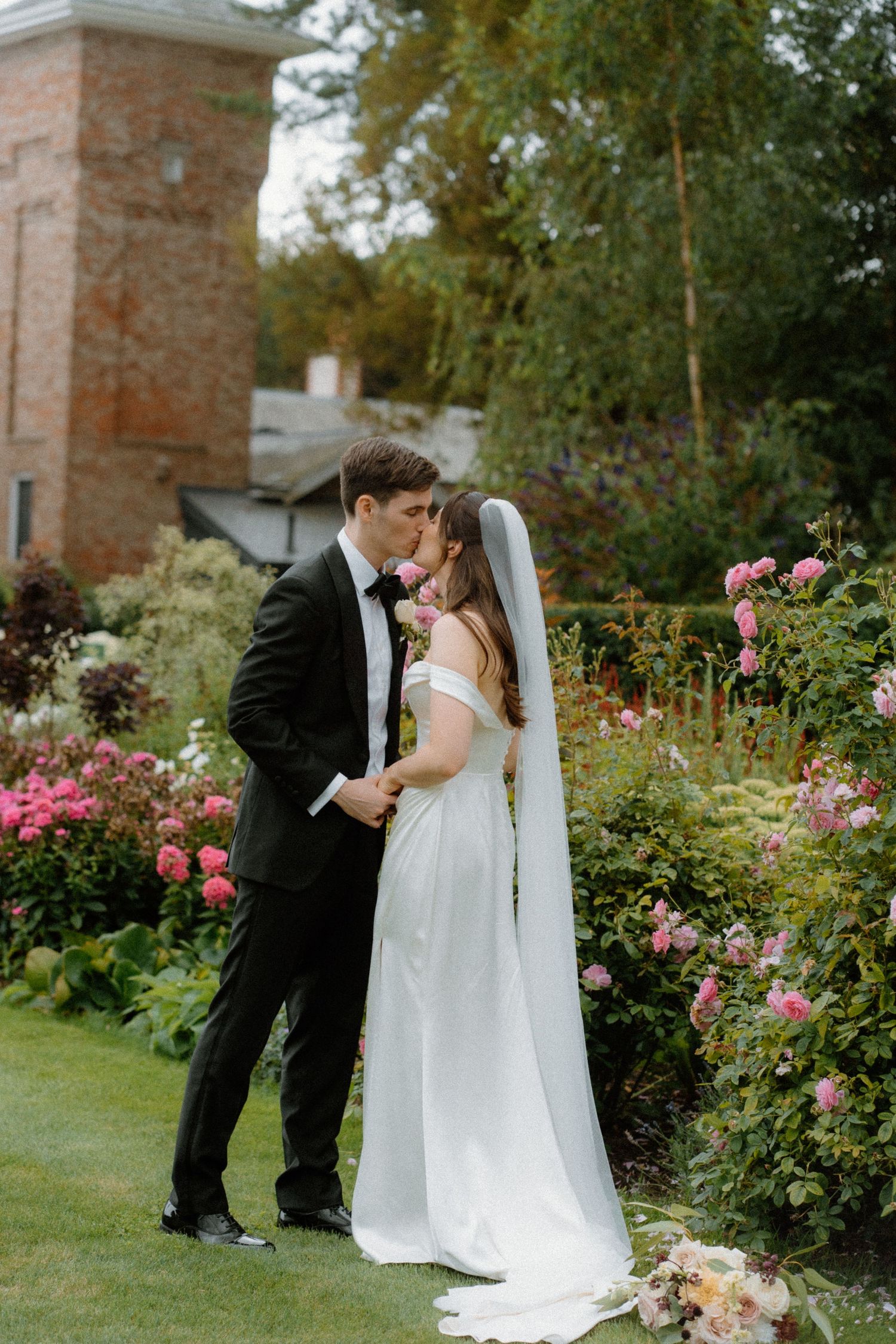 Bride and groom sharing a kiss in a lush garden with pink roses, bride wearing a white gown and veil.