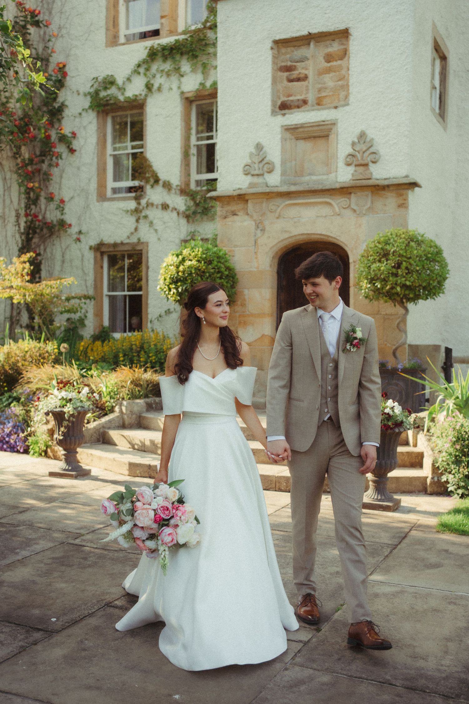 Bride and groom holding hands and smiling outside elegant stone manor with climbing roses.