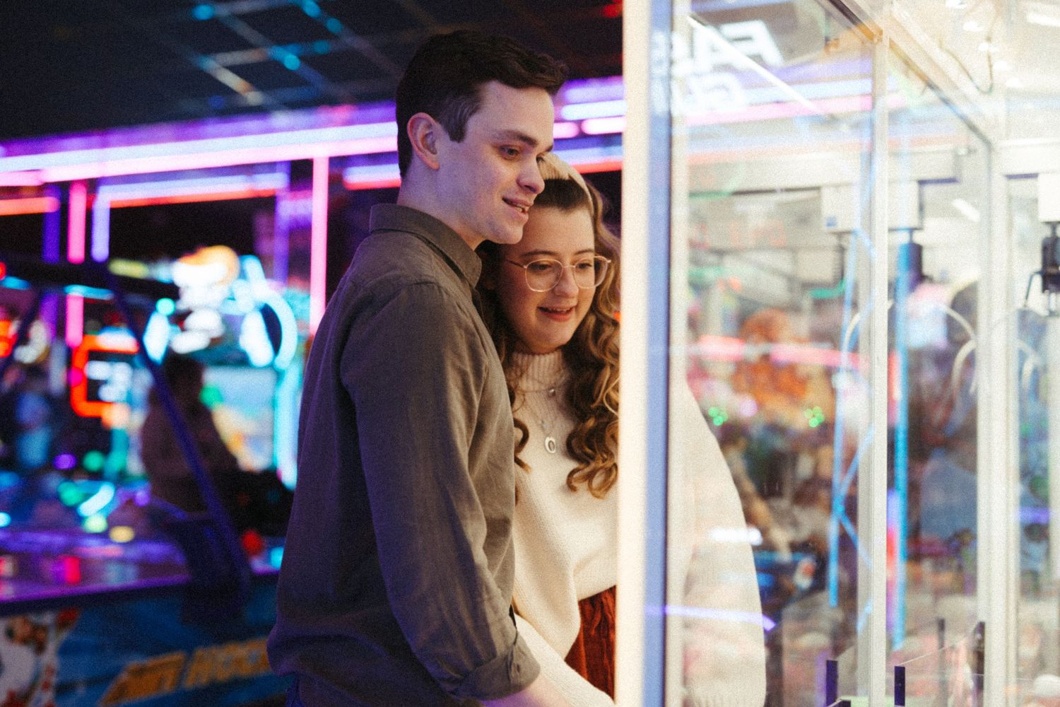 Couple smiling together at an arcade claw machine surrounded by vibrant neon lights.
