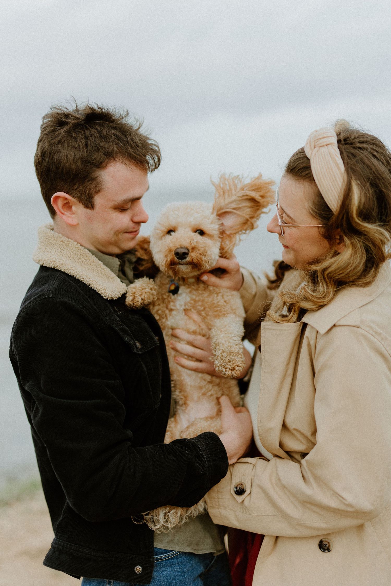 Couple holding fluffy golden doodle dog on windy beach, both smiling at their adorable pet together.