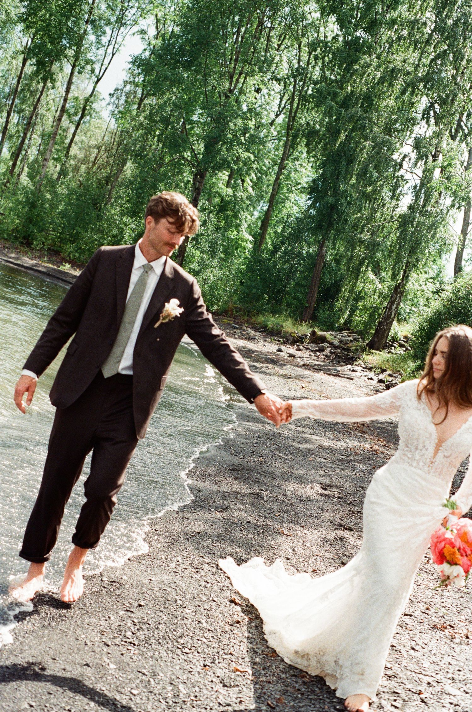 Bride and groom holding hands walking barefoot along a scenic riverbank surrounded by trees.