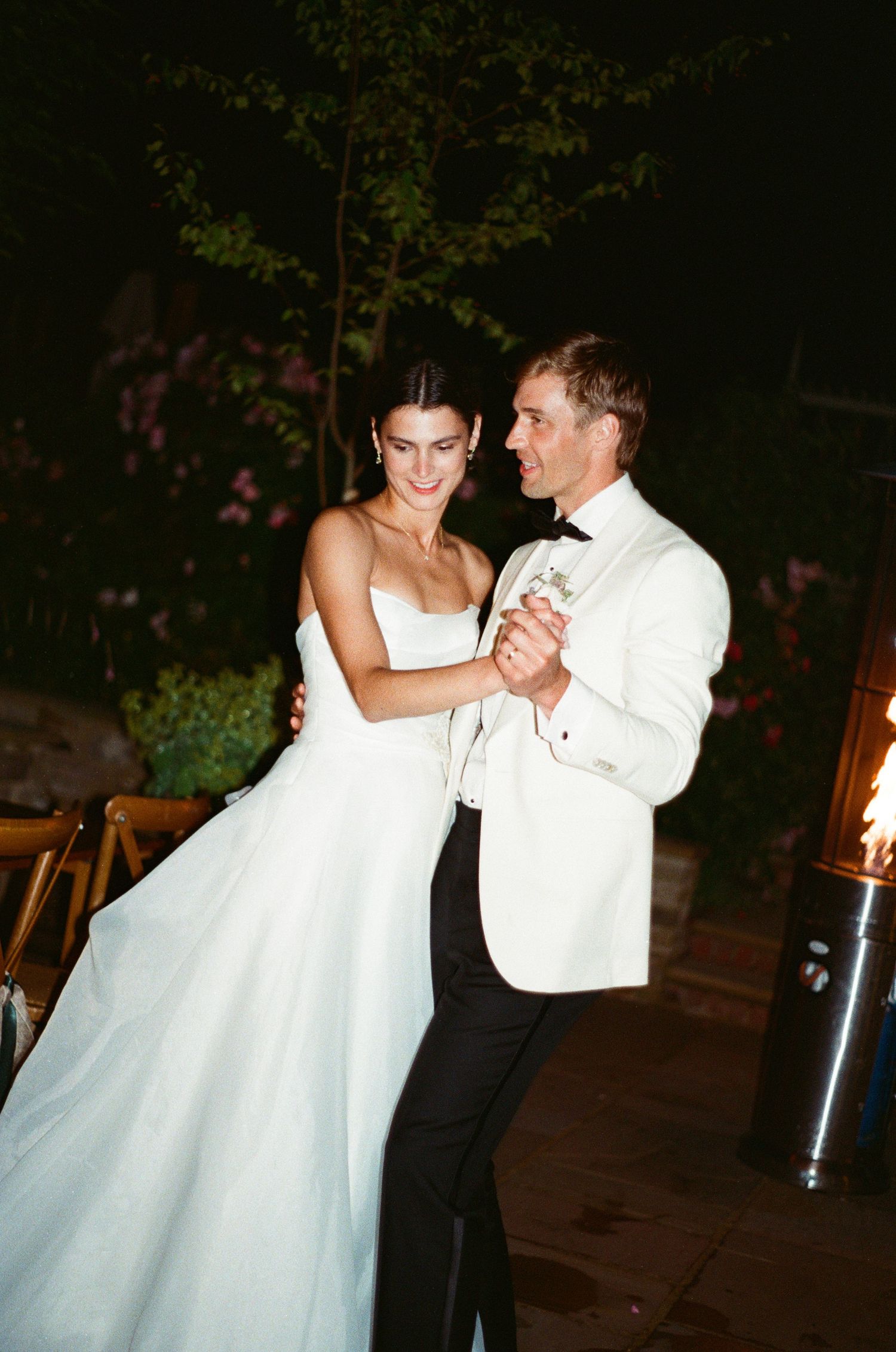 Bride and groom share their first dance outdoors at night surrounded by blooming flowers and fire.