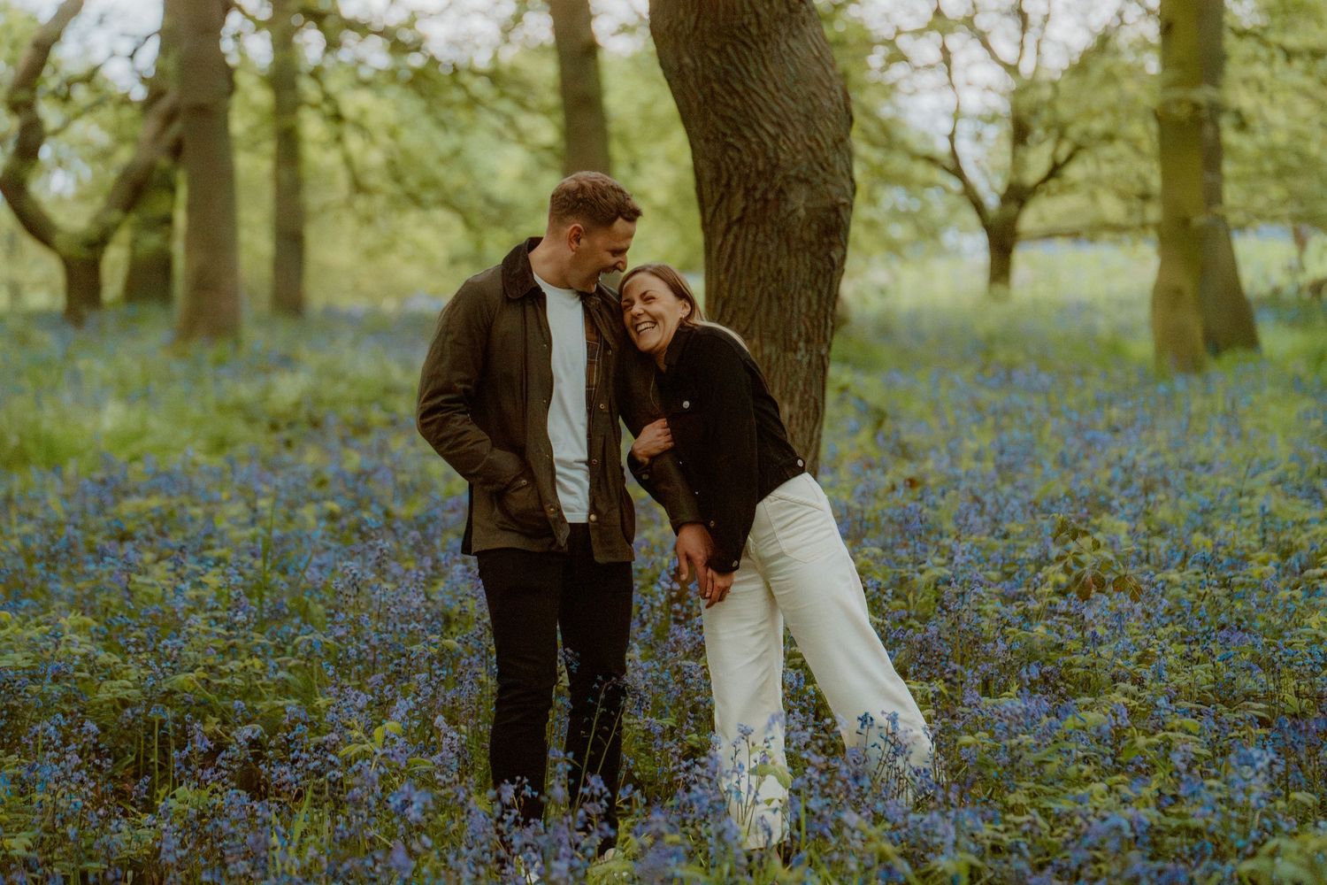 Laughing couple holding hands among vibrant bluebells in a sunlit woodland.