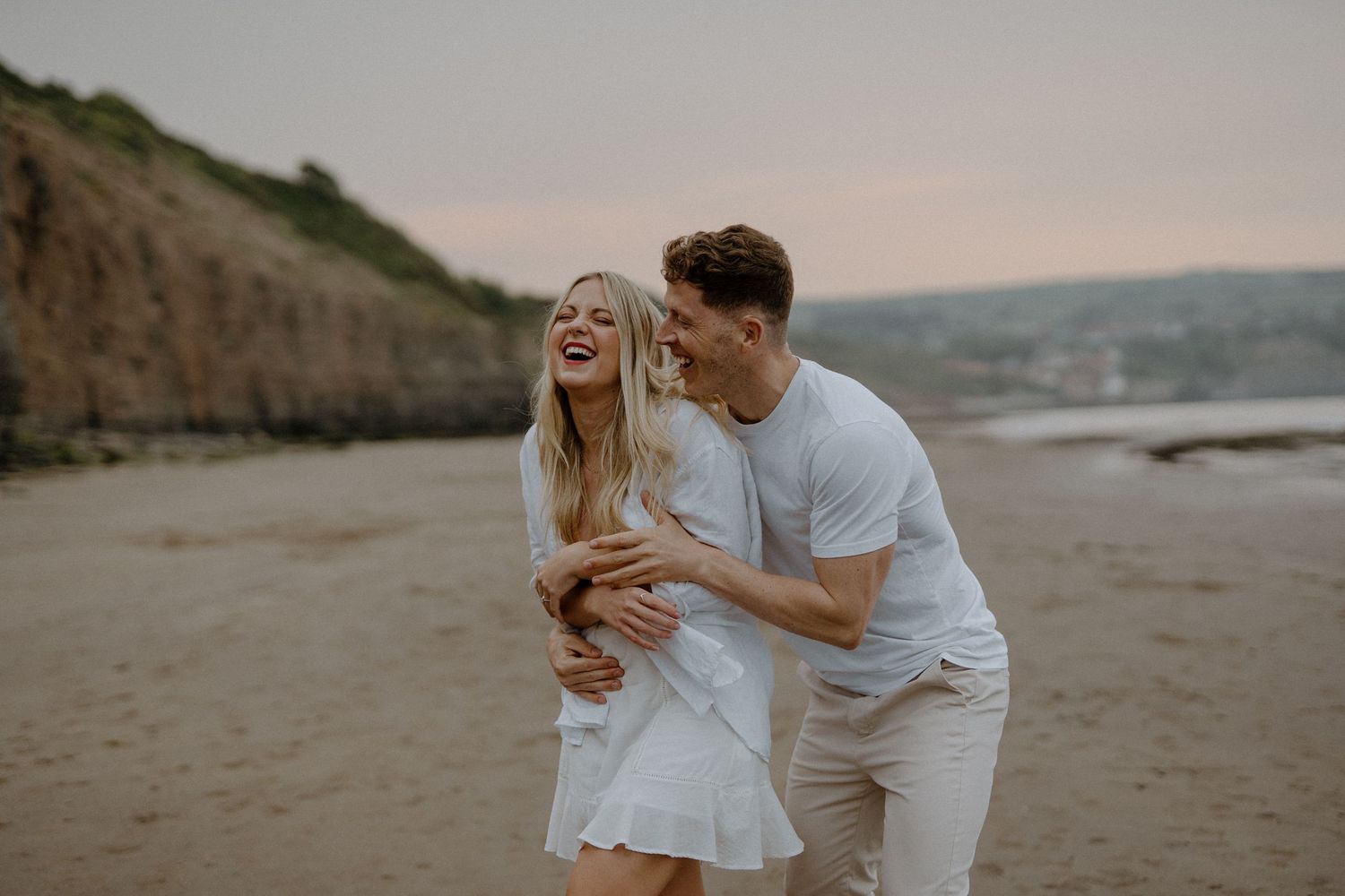 Laughing couple embracing playfully on a sandy beach at sunset with cliffs behind.