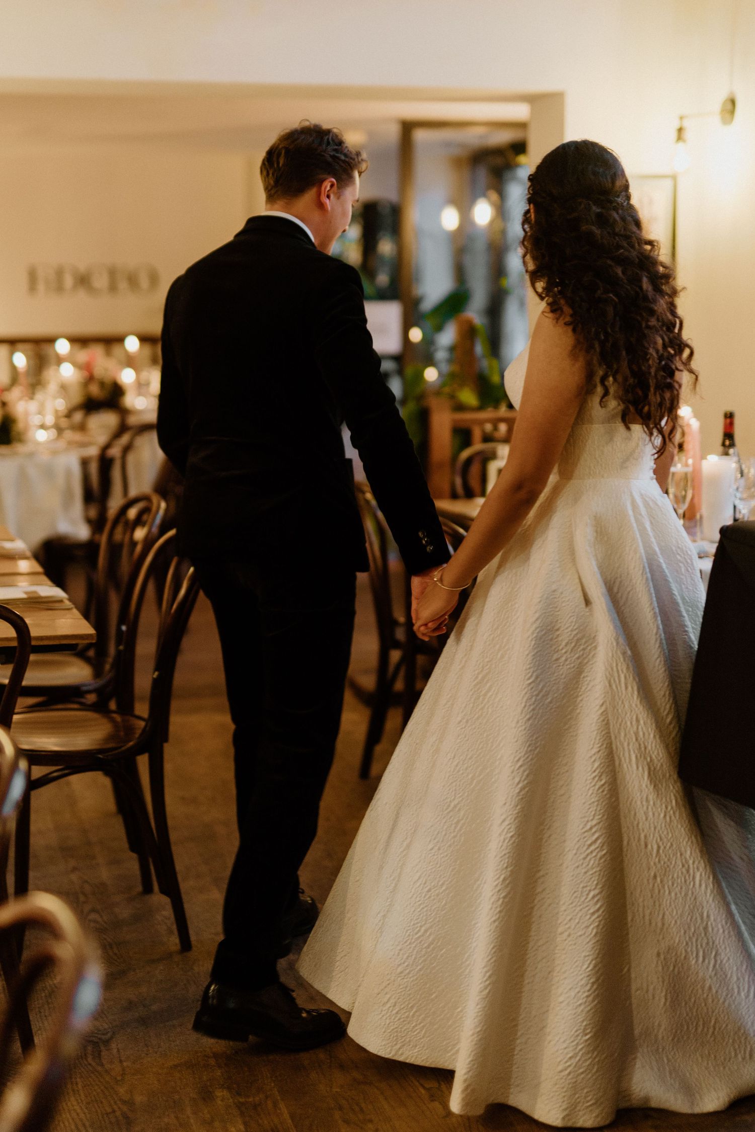 Bride and groom holding hands from behind walking through their candlelit wedding reception venue.