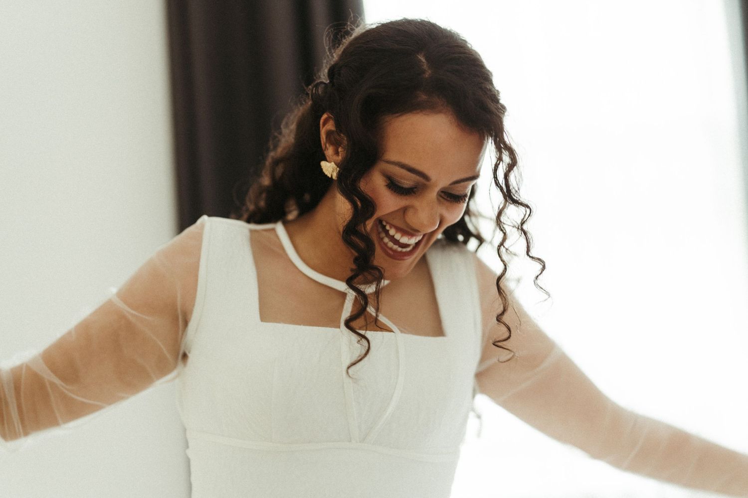Joyful bride in white square-neck wedding dress with sheer sleeves laughing and spreading her arms wide.