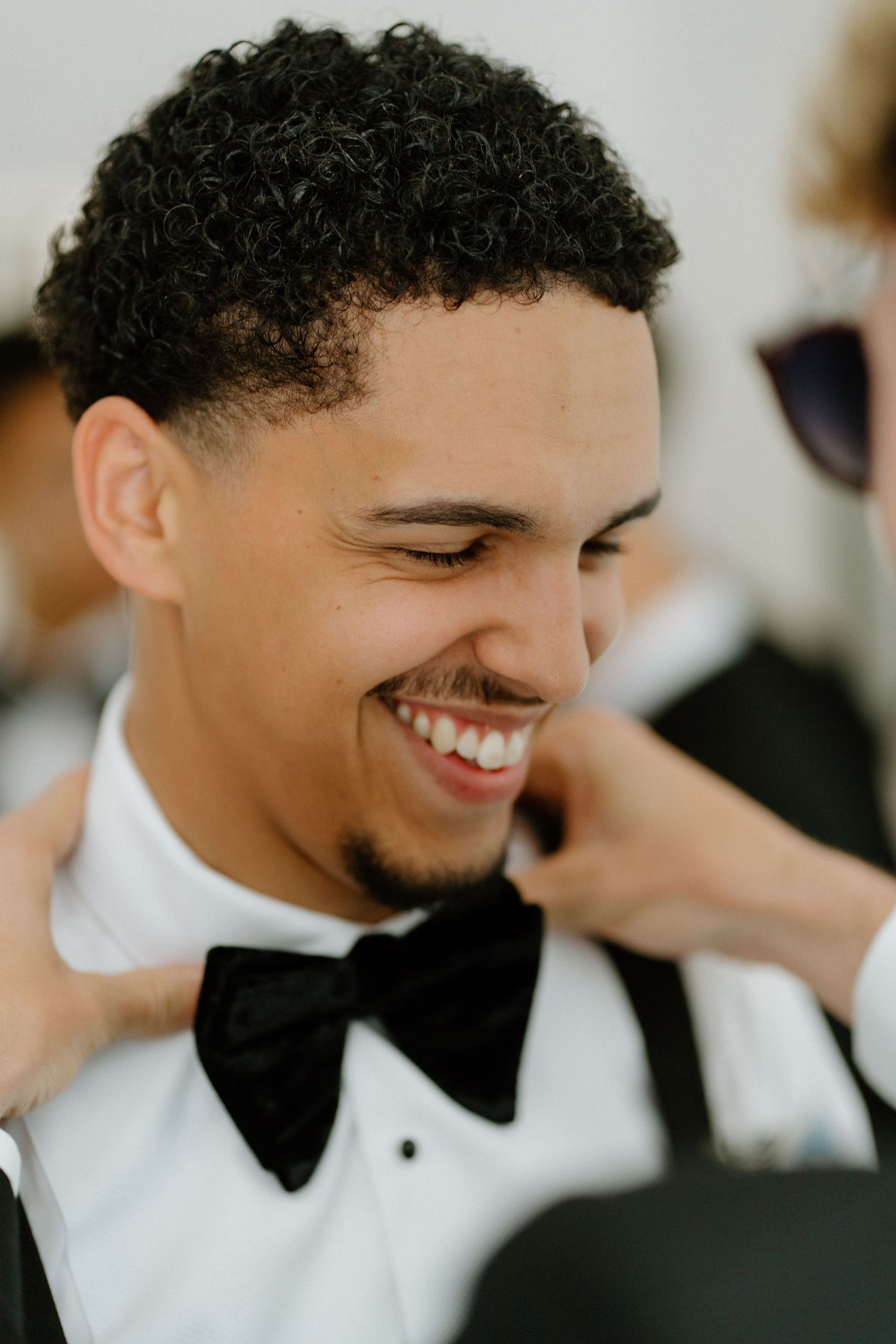 Groom in white tuxedo shirt laughing joyfully as someone adjusts his black bow tie before the wedding.