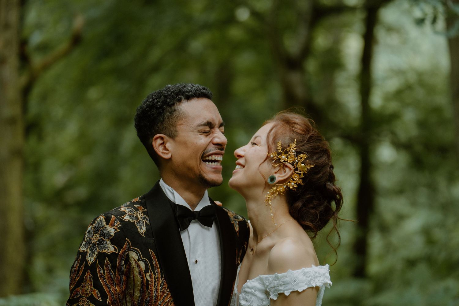 Joyful bride and groom laughing together in a woodland setting, groom wearing gold batik blazer.
