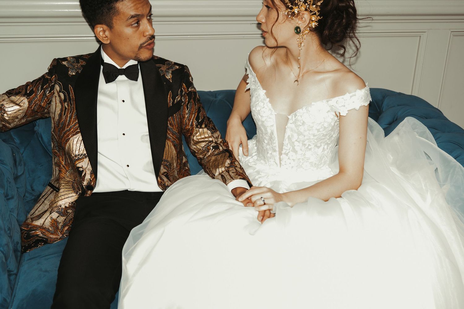 Bride in white lace off-shoulder gown and groom in gold patterned tuxedo sitting on blue velvet sofa.