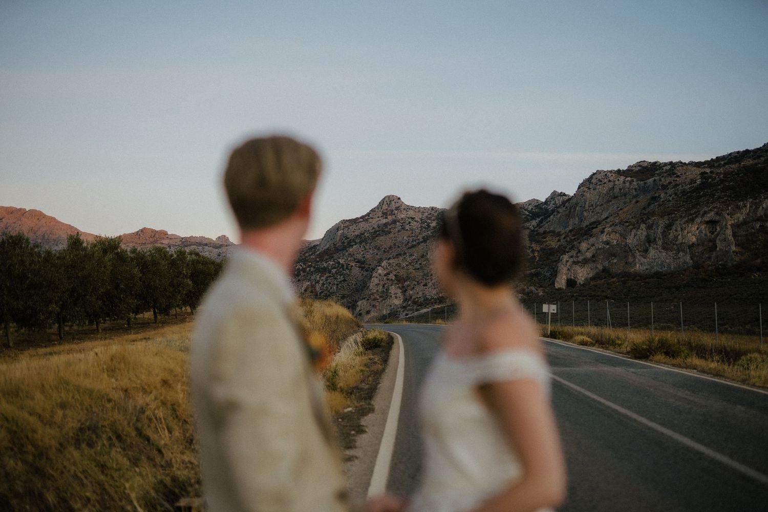 Newlywed couple standing on rural road gazing at stunning rocky mountain landscape at dusk.