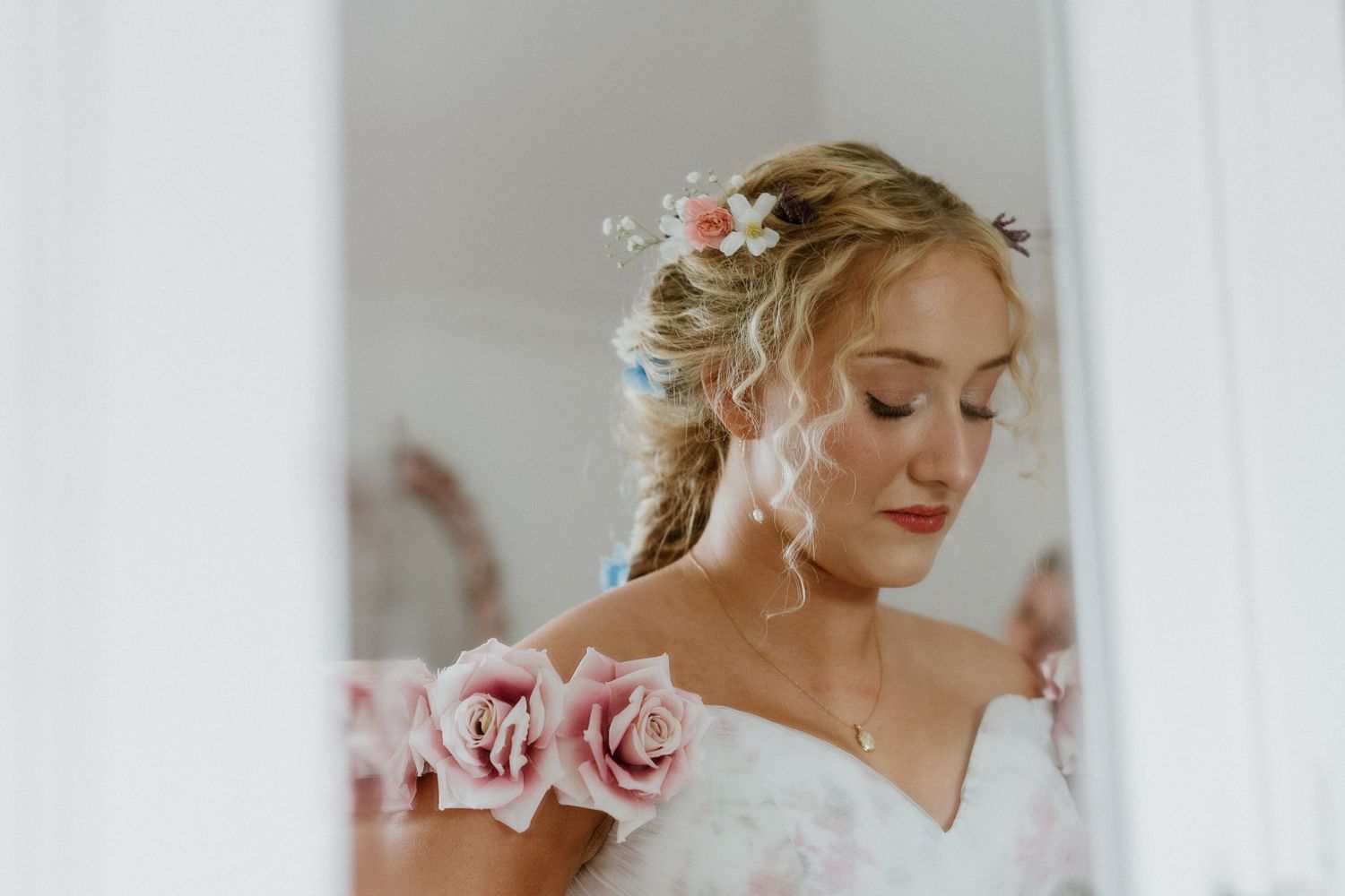 Soft portrait of a bride with pink roses wearing delicate off-shoulder wedding dress and elegant hairstyle.