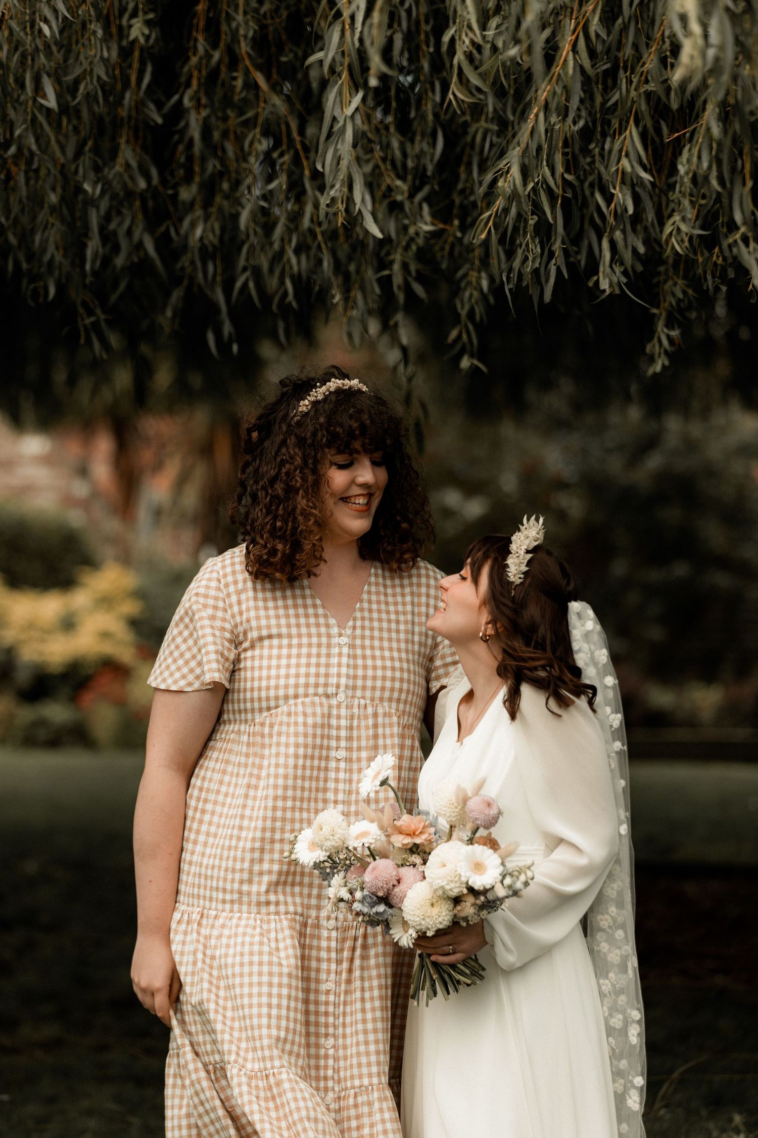 Two wedding attendees share a joyful moment under hanging willow branches in a garden setting.