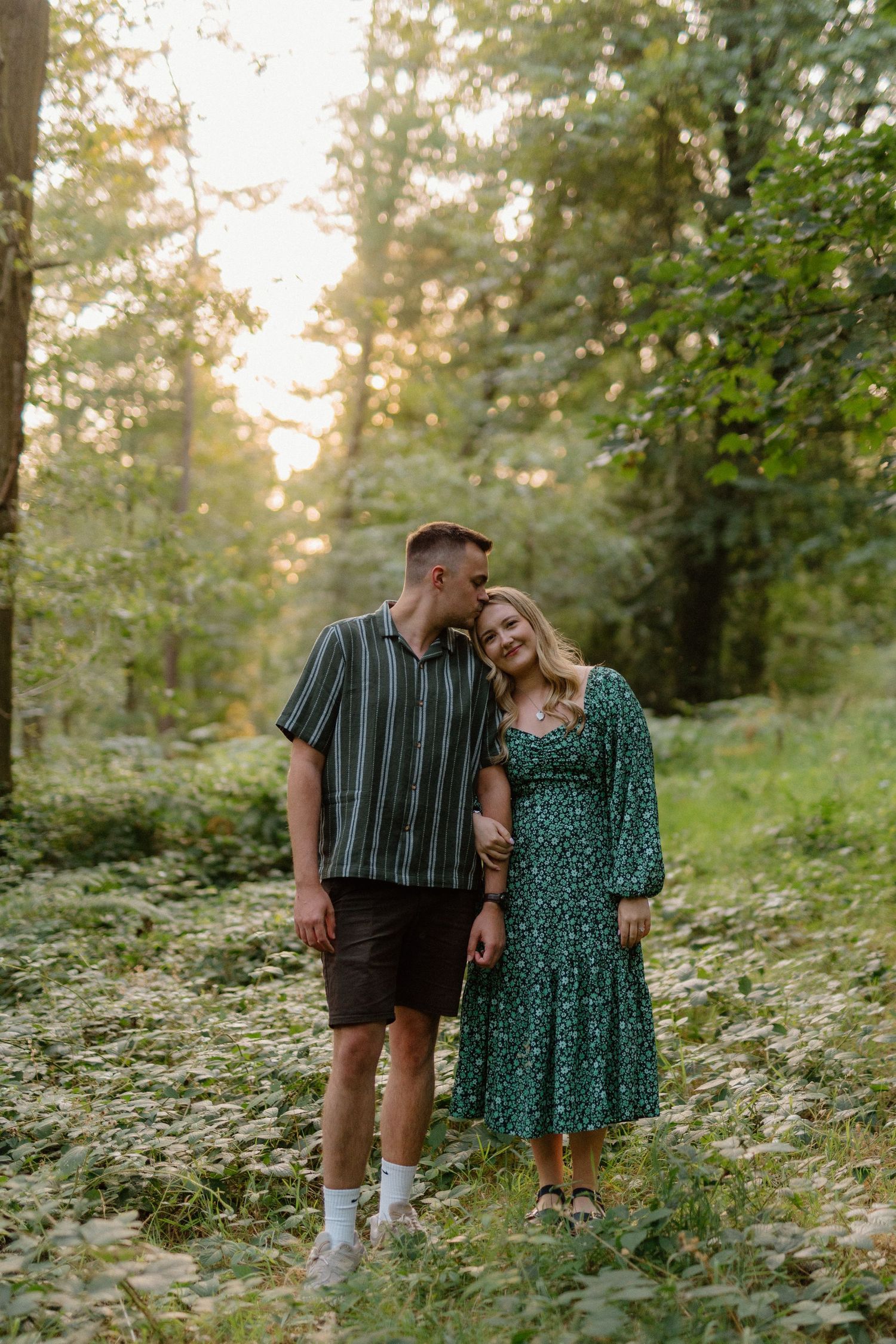 A couple shares an intimate moment on a wooded path during golden hour in summer nature setting.