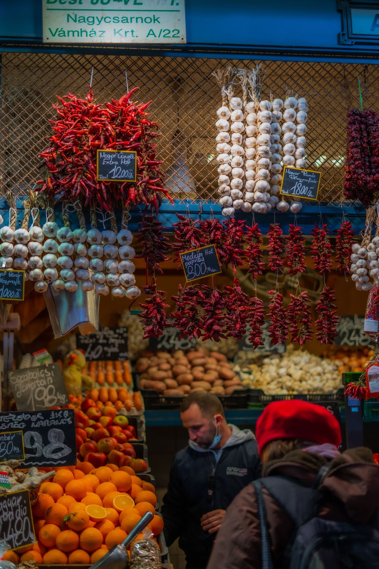 Colorful market stall displays fresh oranges, strings of red chili peppers, and garlic braids at a bustling produce stand.