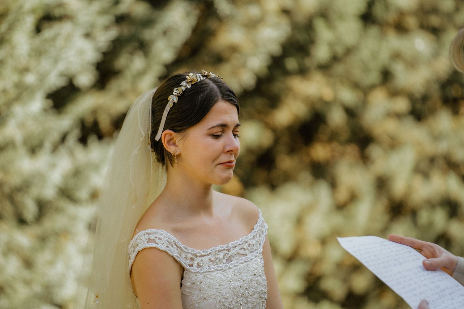 Emotional bride in lace off-shoulder gown and floral headpiece listening to vows outdoors.