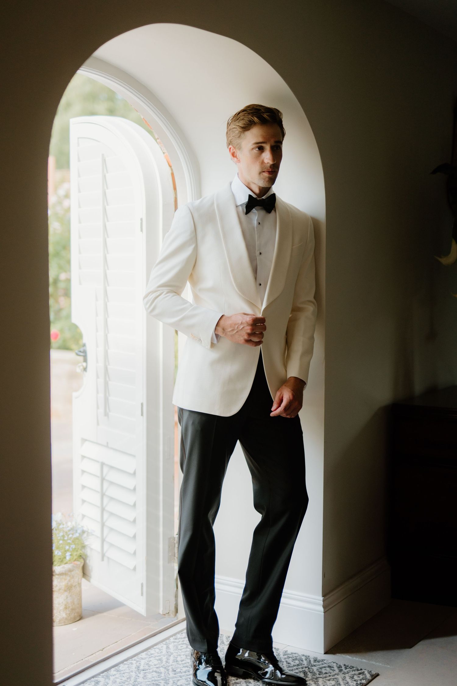Dapper groom in white tuxedo jacket and black bow tie stands in arched doorway before wedding.