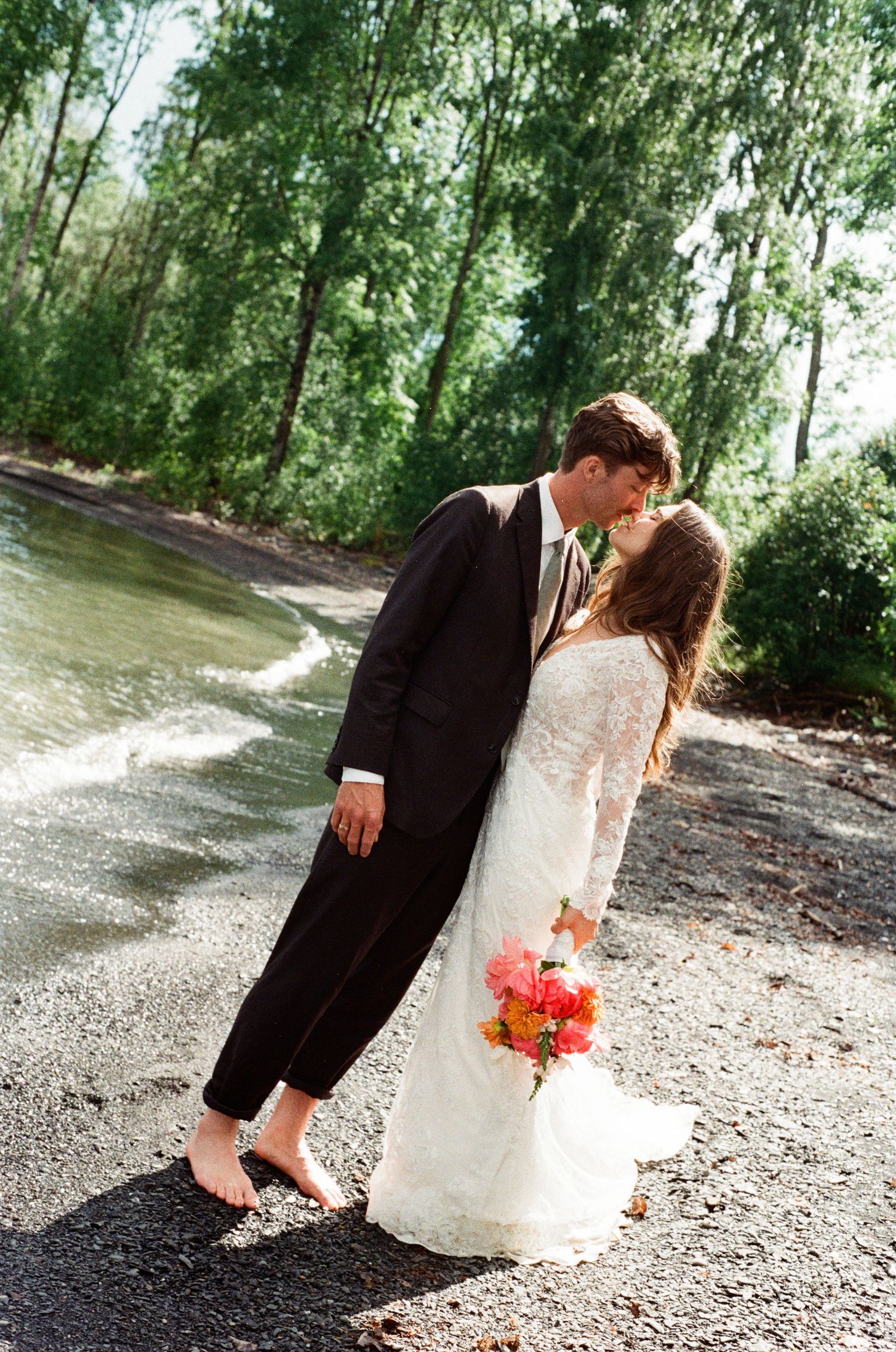 Barefoot bride and groom sharing a kiss on a pebble beach surrounded by lush green trees.