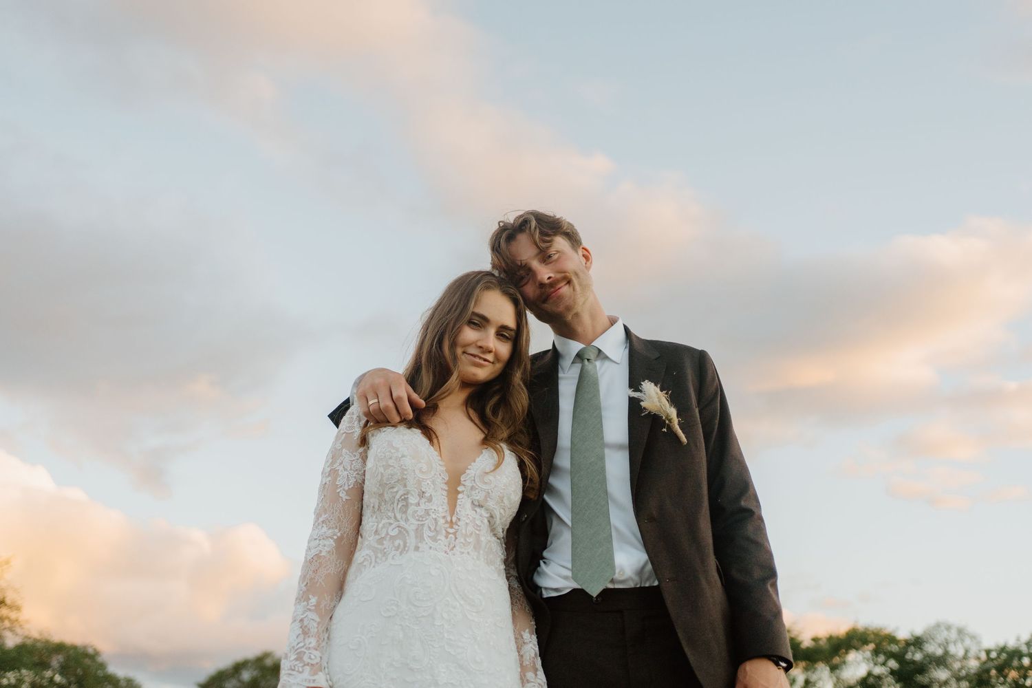 Bride and groom posing together outdoors at sunset with a dramatic cloudy sky backdrop.