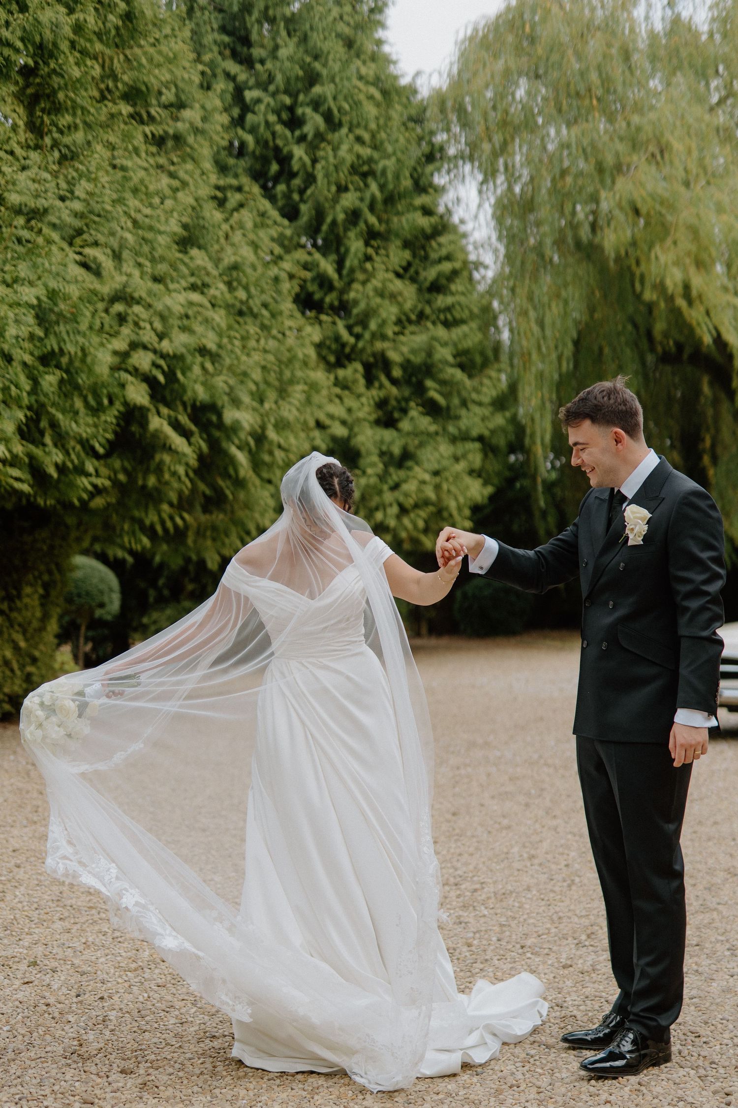 Bride and groom hold hands outdoors as her veil flows in the wind, surrounded by lush green trees.