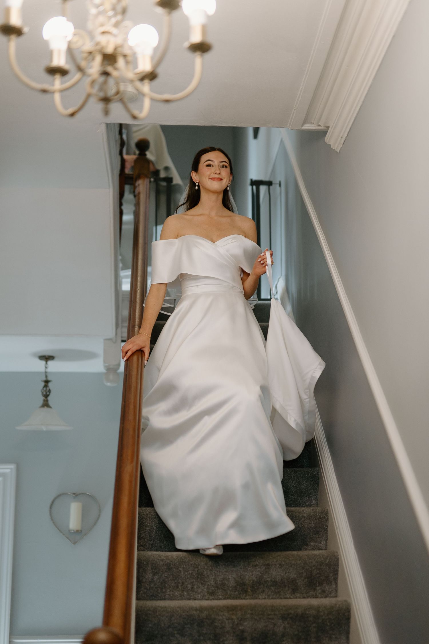 Beautiful bride in off-shoulder white satin gown descending staircase before wedding ceremony.