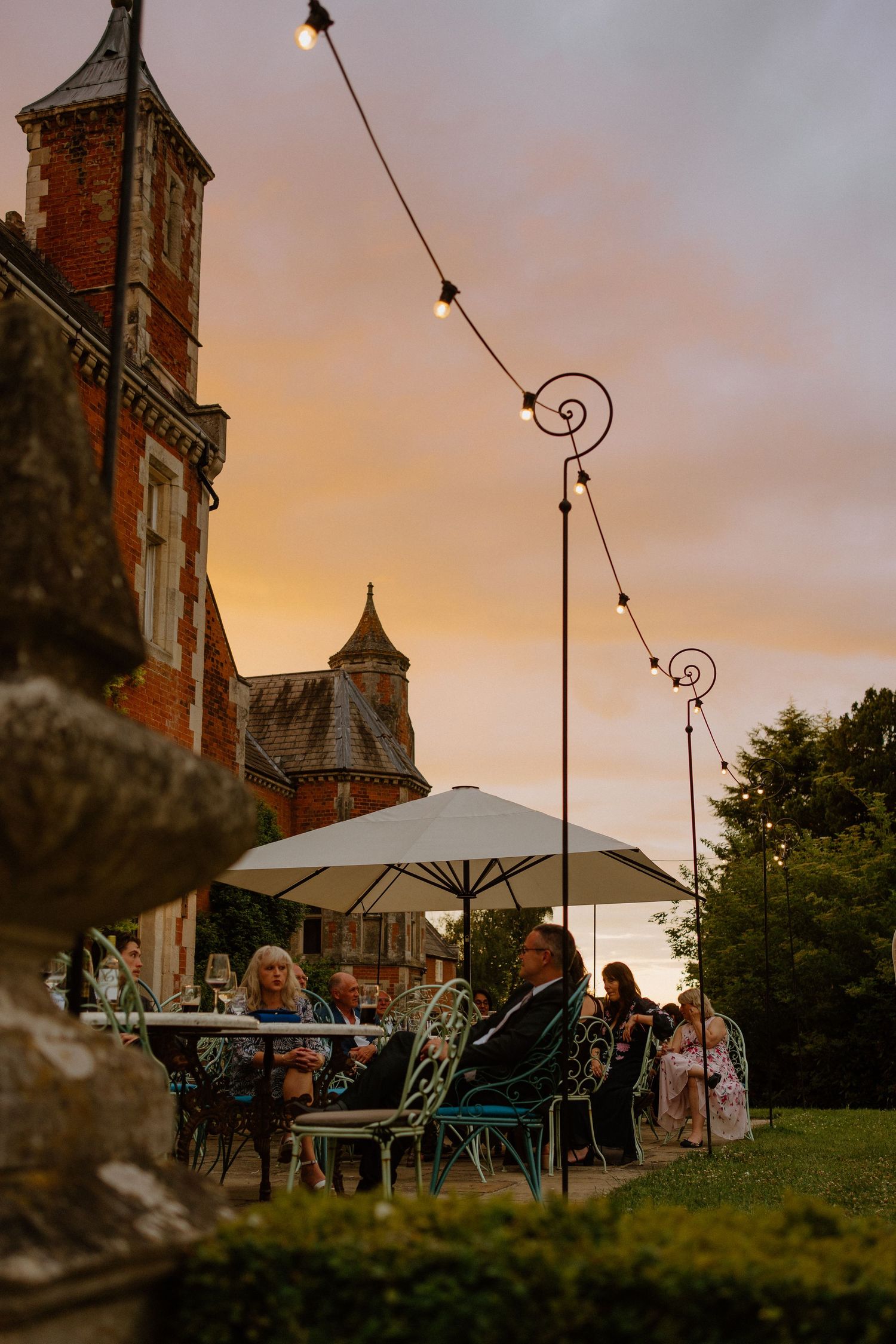 Wedding guests dining outdoors at sunset beside historic red brick manor with festoon lights overhead.
