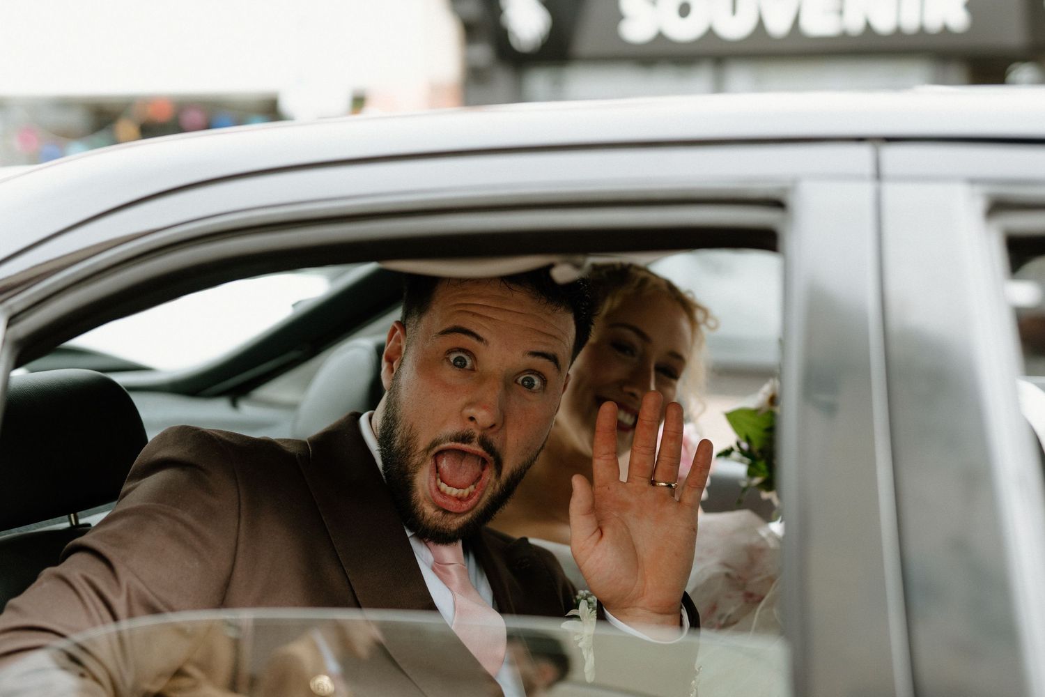 Excited newlyweds wave and smile from a car window after their wedding ceremony.