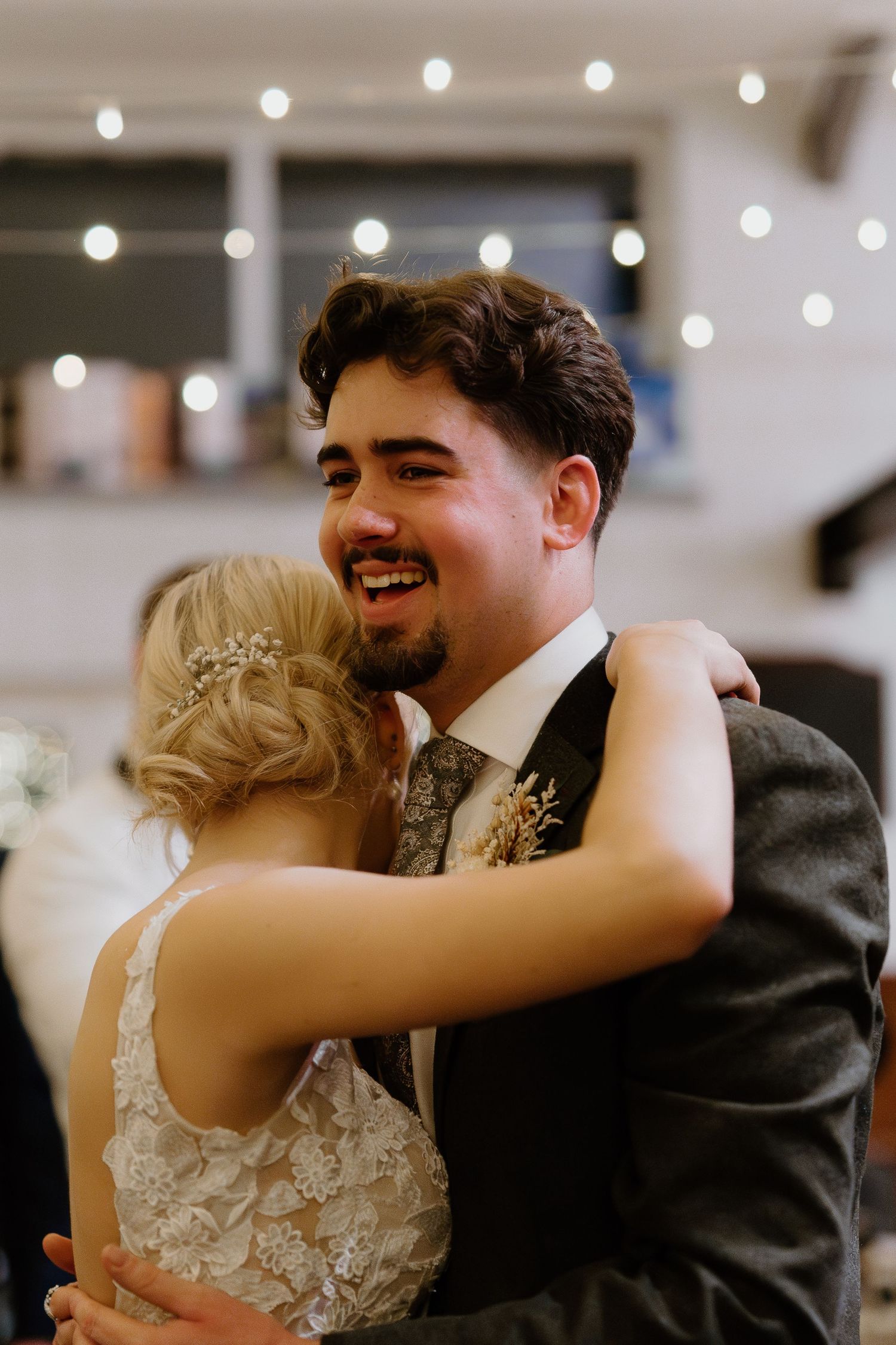 Smiling groom in grey suit hugging bride in lace wedding dress during first dance at romantic wedding reception.