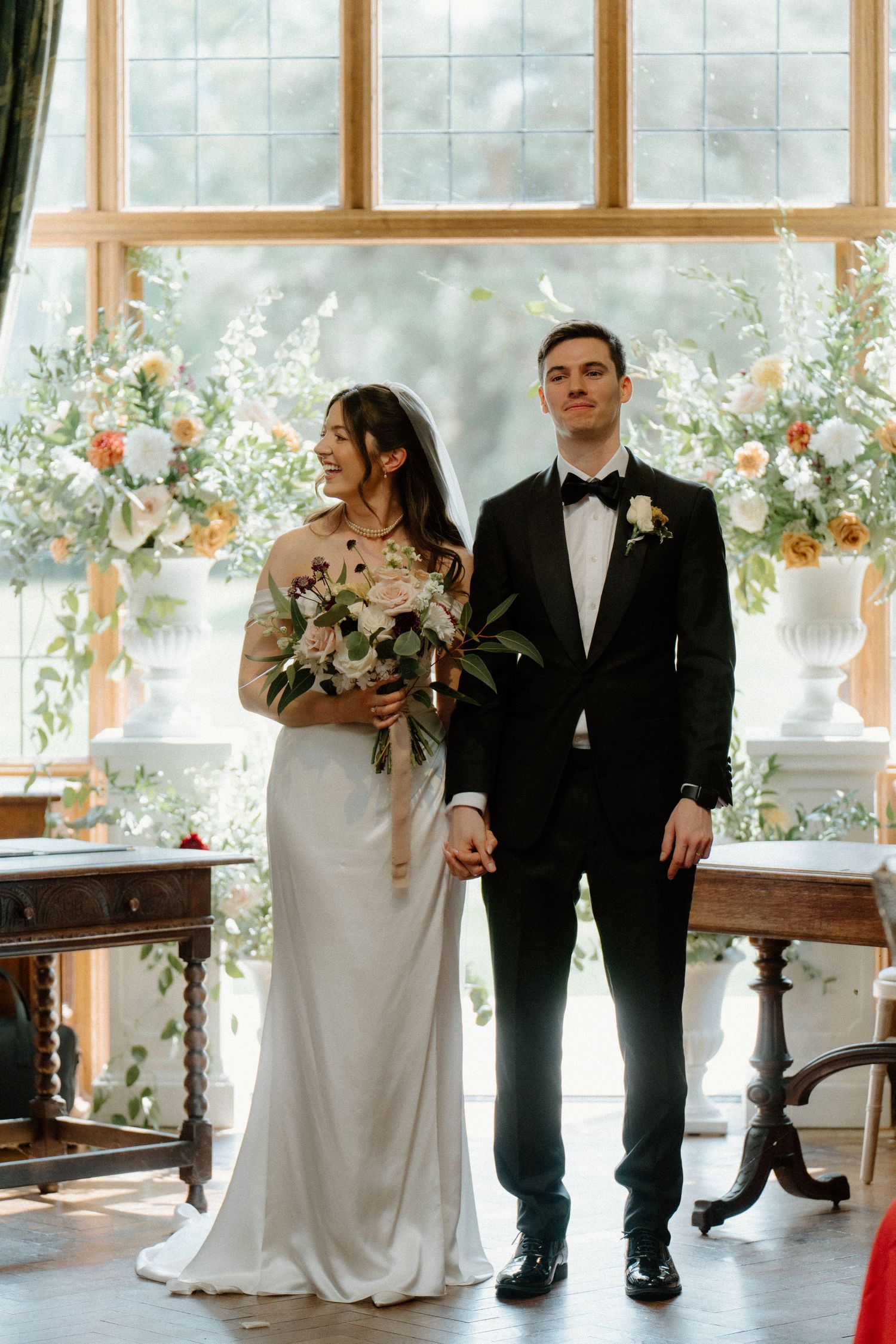 Bride holding bouquet and groom in black tuxedo stand before floral arch at wedding ceremony.