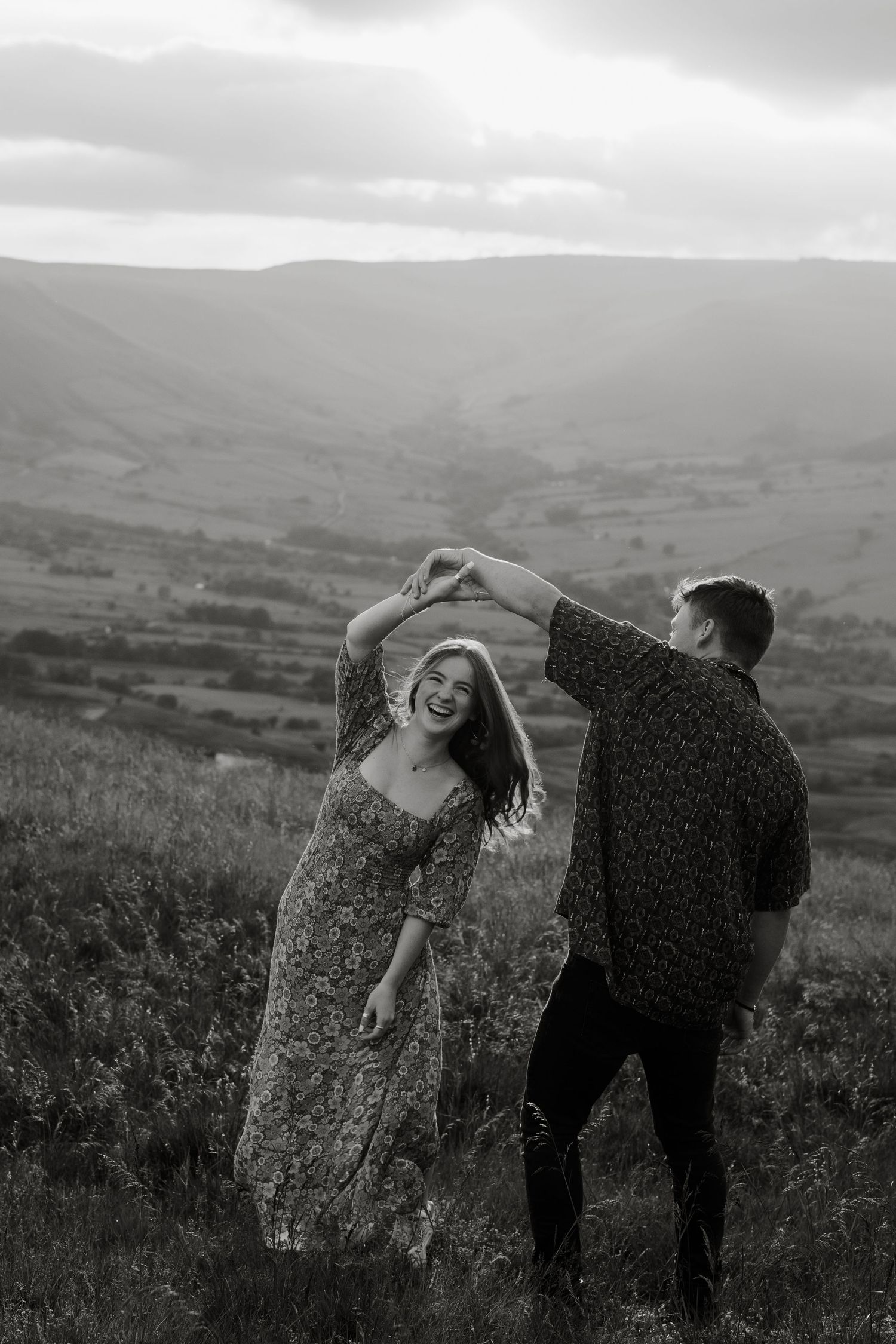 Black and white photo of joyful couple dancing on a hilltop with mountain views.