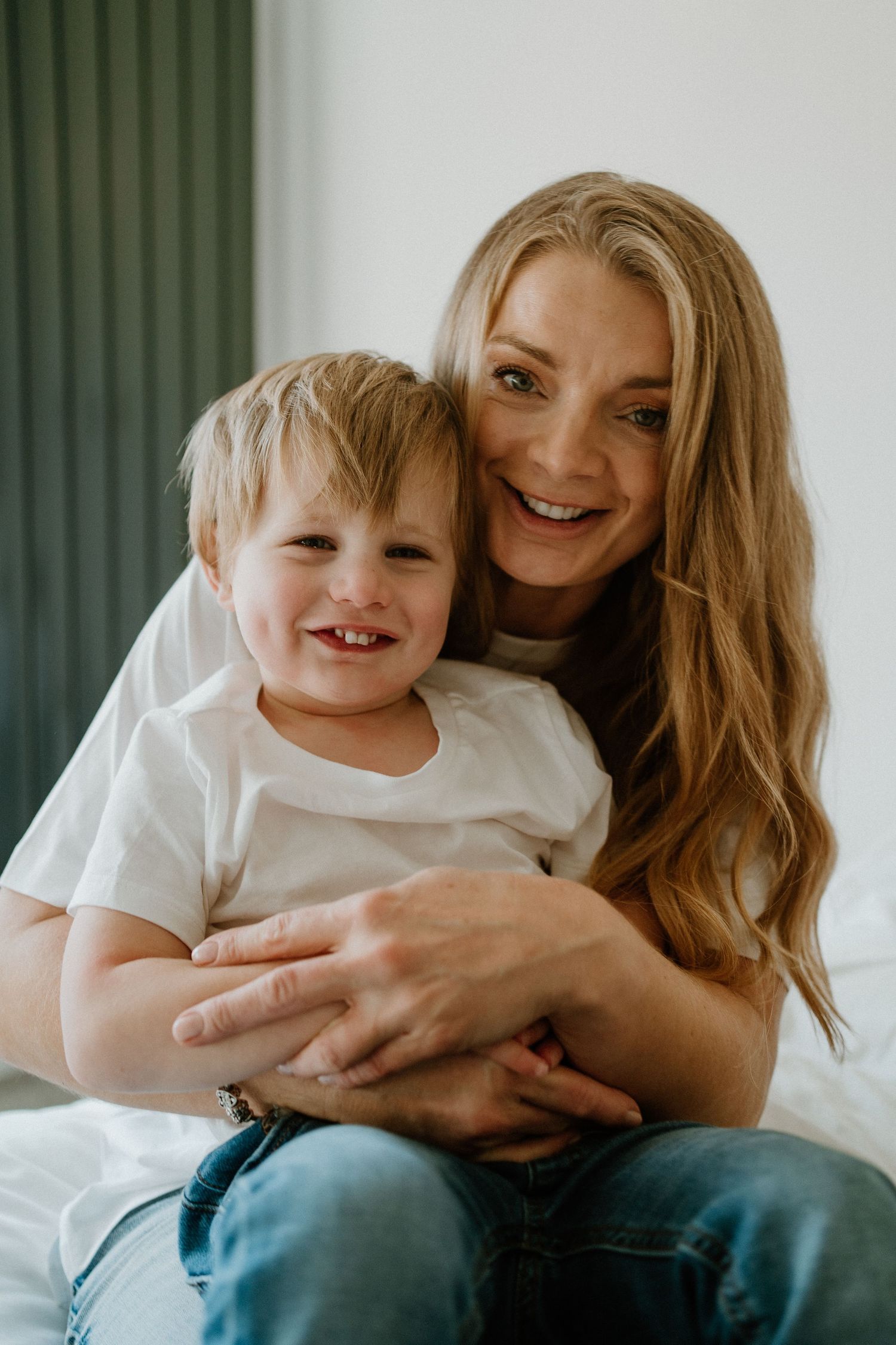 Smiling blonde mother hugging her young son while sitting together on a white bed.