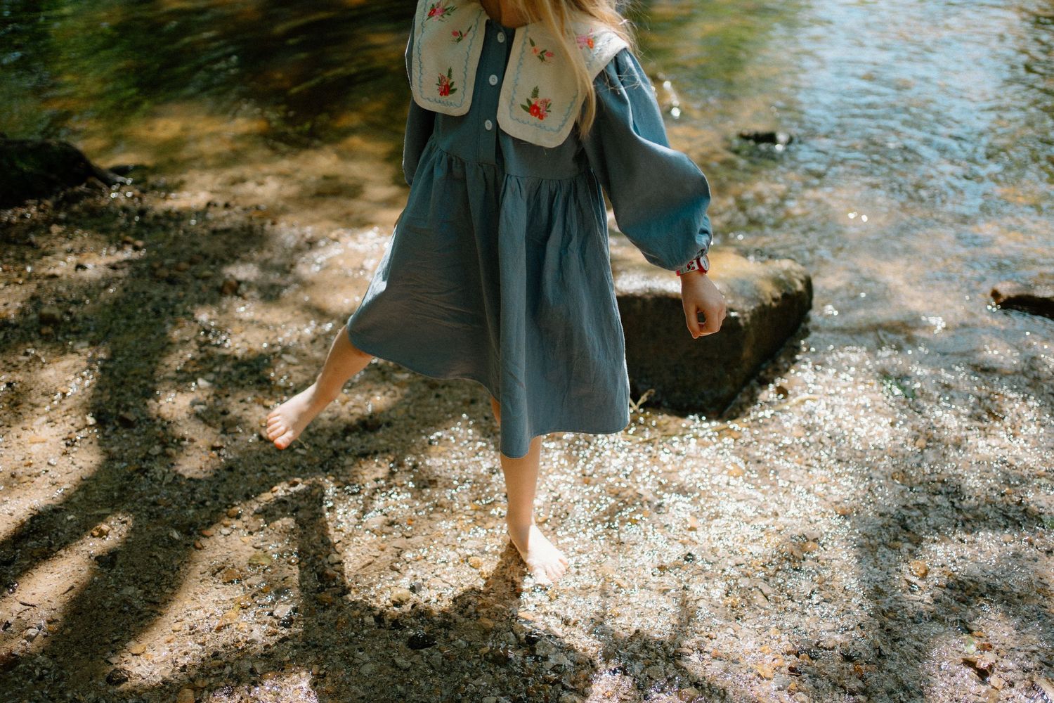 A barefoot child in a blue dress and backpack walks along a sunlit rocky riverbank.