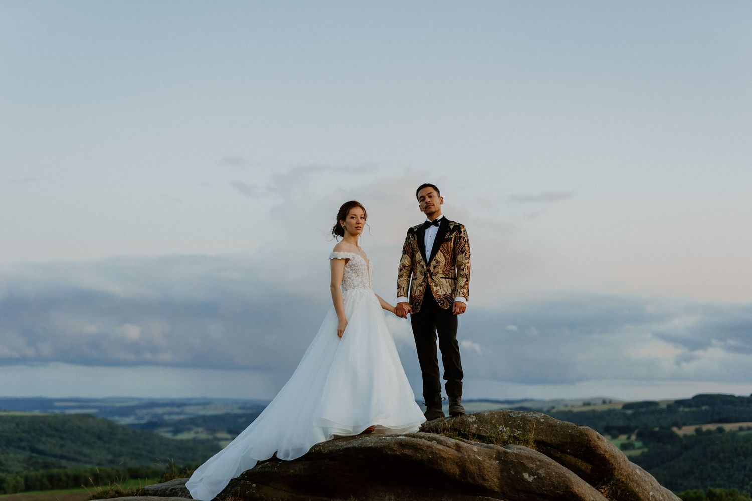 Bride and groom holding hands on rocky hilltop at sunset with panoramic landscape views.