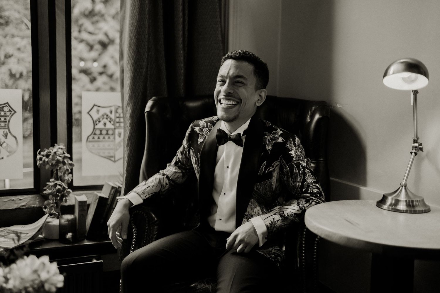 Groom in ornate gold blazer laughs while seated in leather chair, black and white photo.