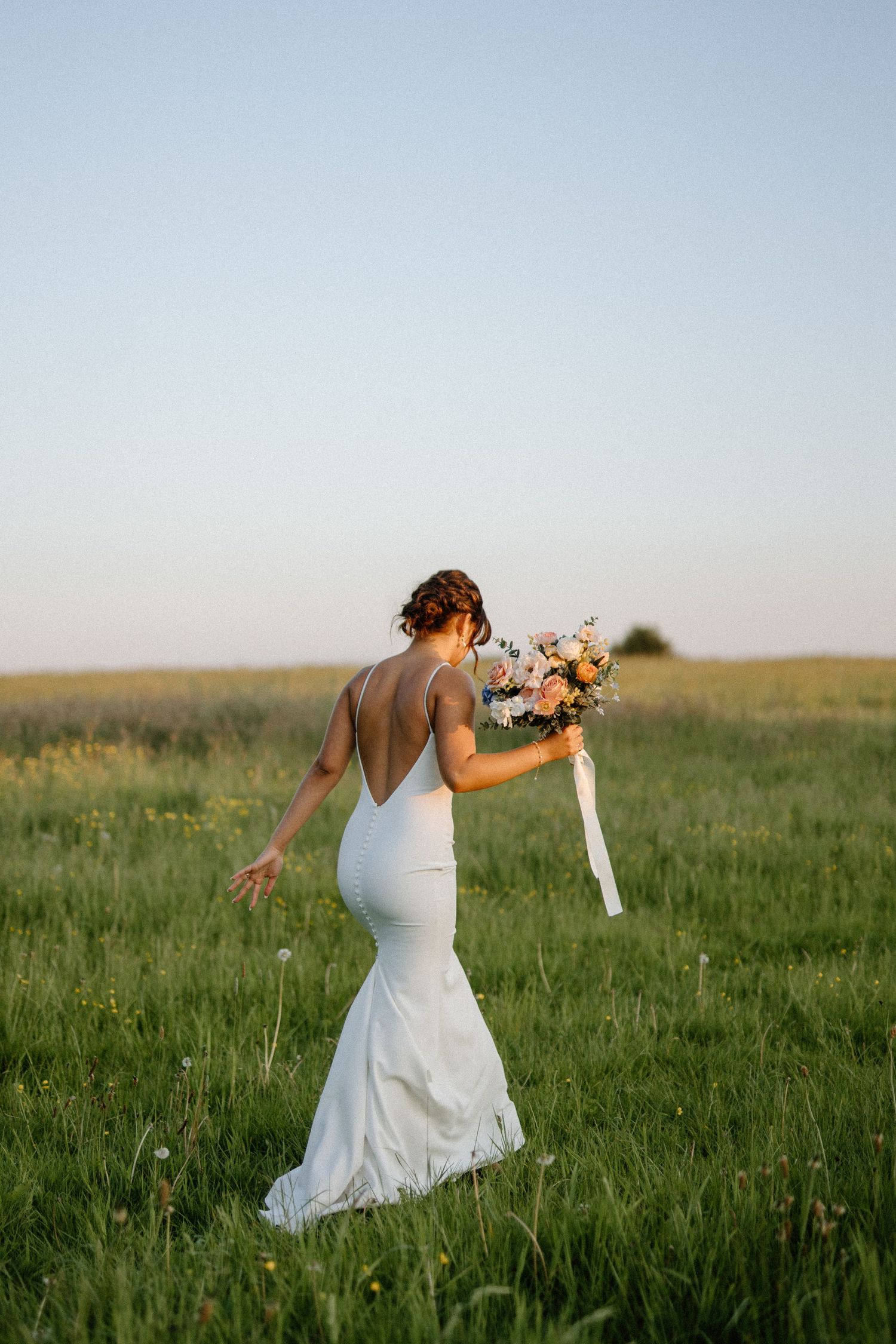 Bride in backless white mermaid gown holding floral bouquet while walking through green meadow at sunset.