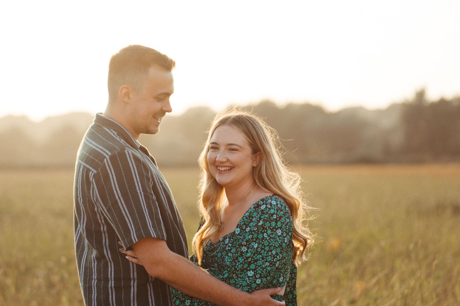 A couple embraces in a sunlit field during golden hour, sharing a joyful moment together in the warm evening light.