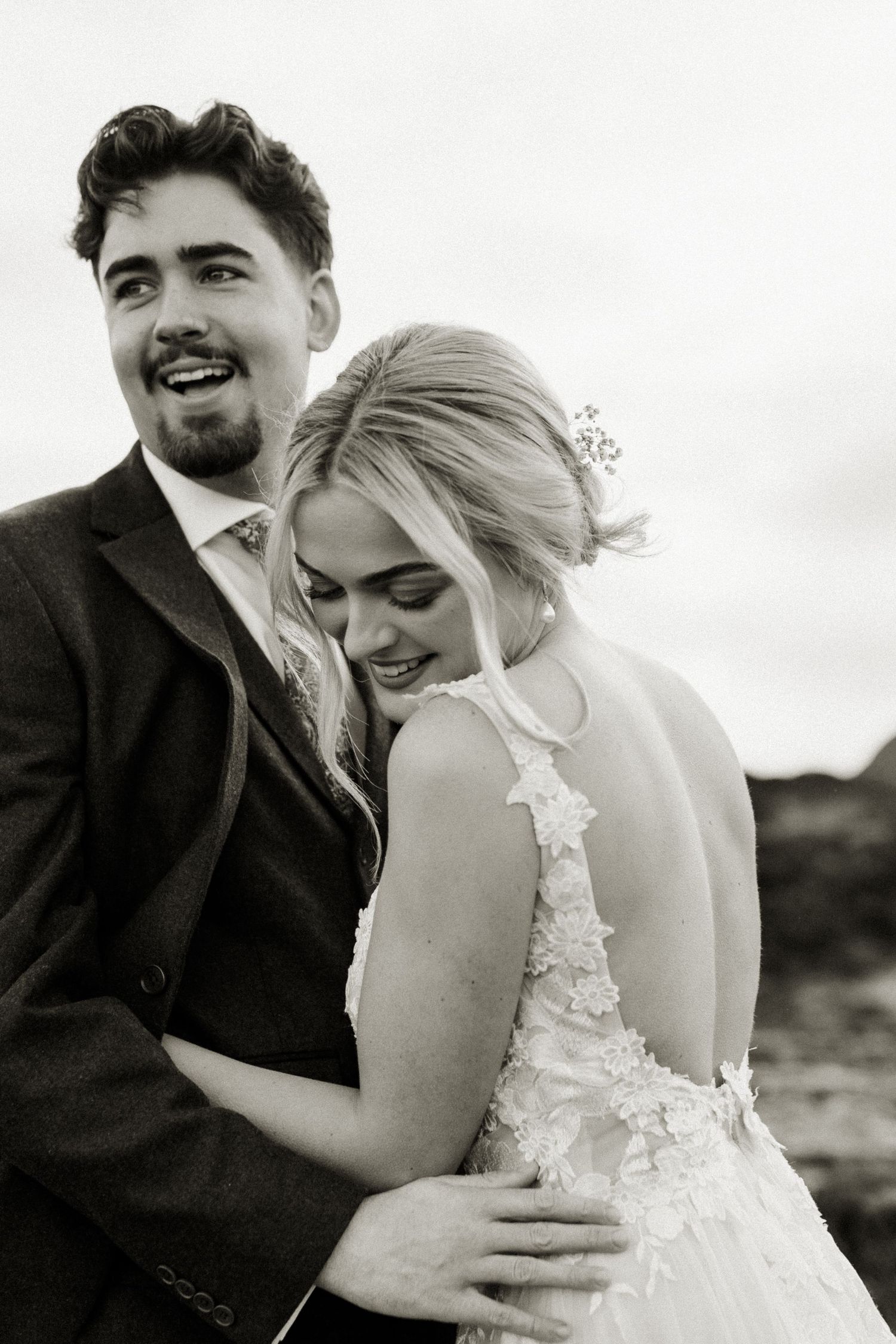 Black and white portrait of laughing groom embracing smiling bride showing elegant open-back floral lace wedding gown.
