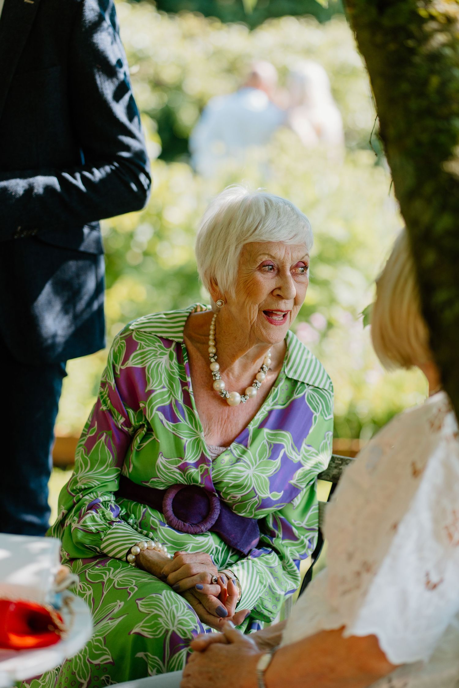 Elegant elderly woman in green floral dress wearing pearl necklace conversing at outdoor wedding.