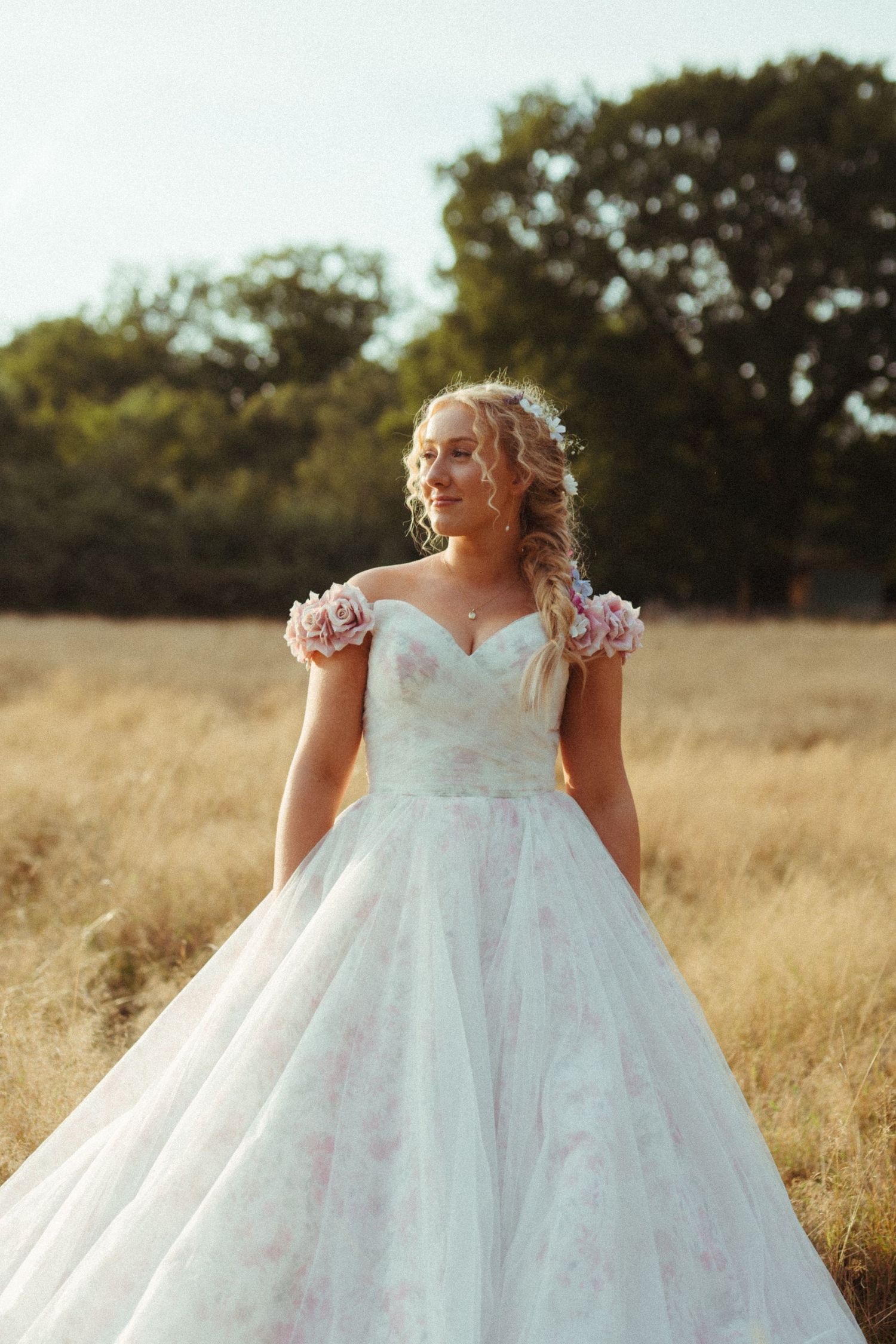 Beautiful bride in off-shoulder white gown with pink floral details stands in golden field at sunset.