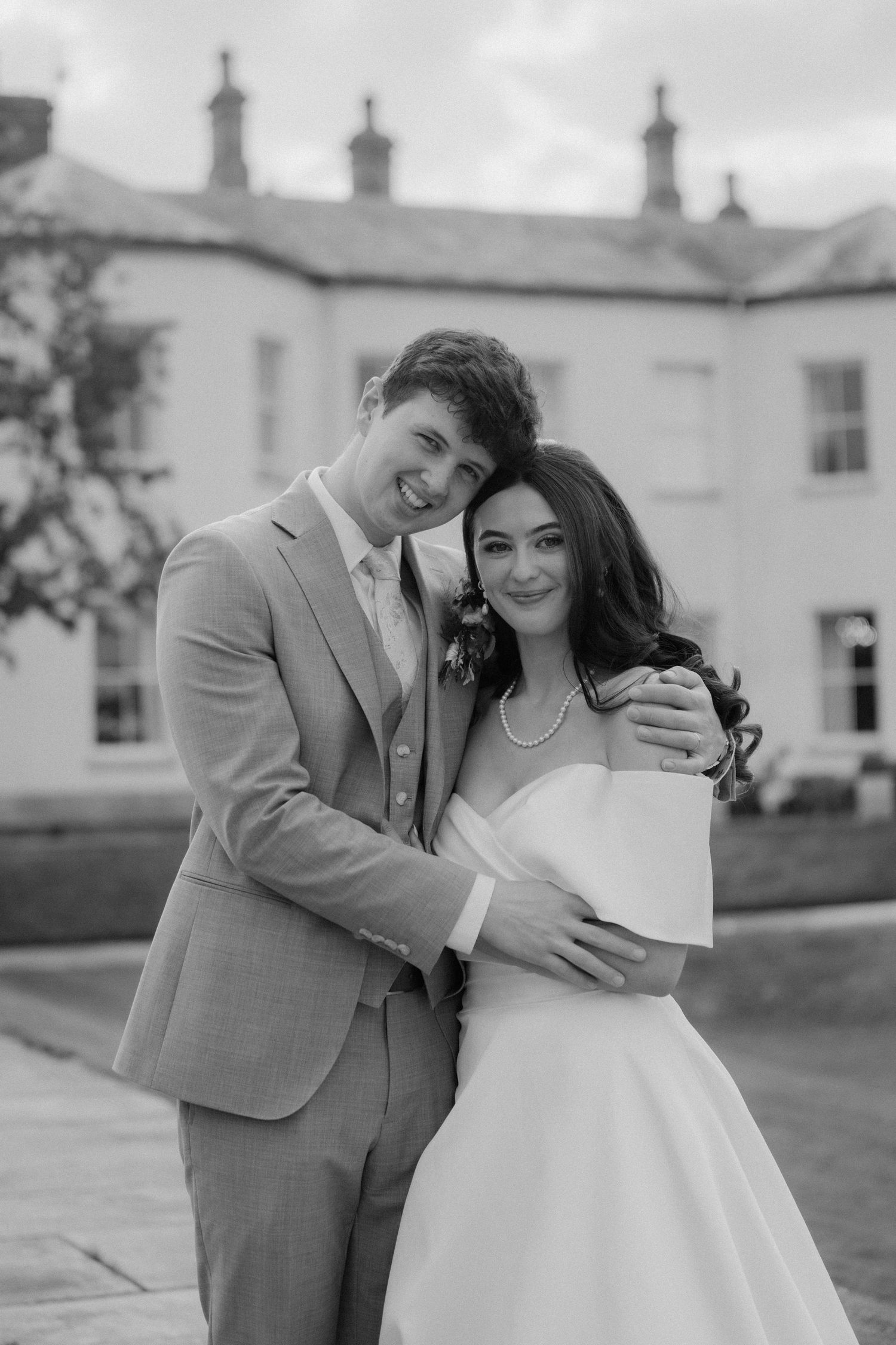 Black and white portrait of a groom embracing his bride in front of an elegant manor house on their wedding day.