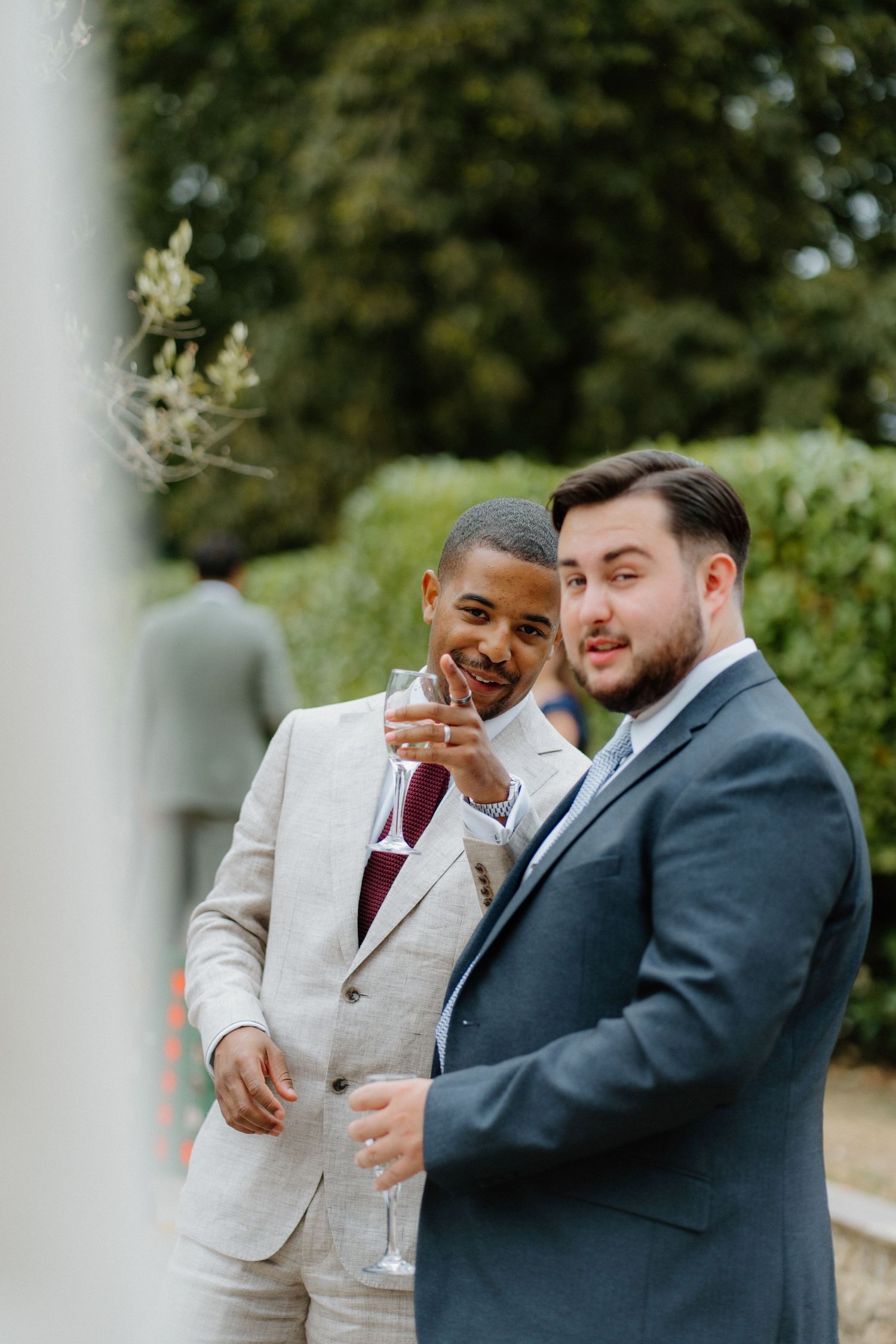 Two male wedding guests smile and clink champagne glasses outdoors at garden wedding reception.