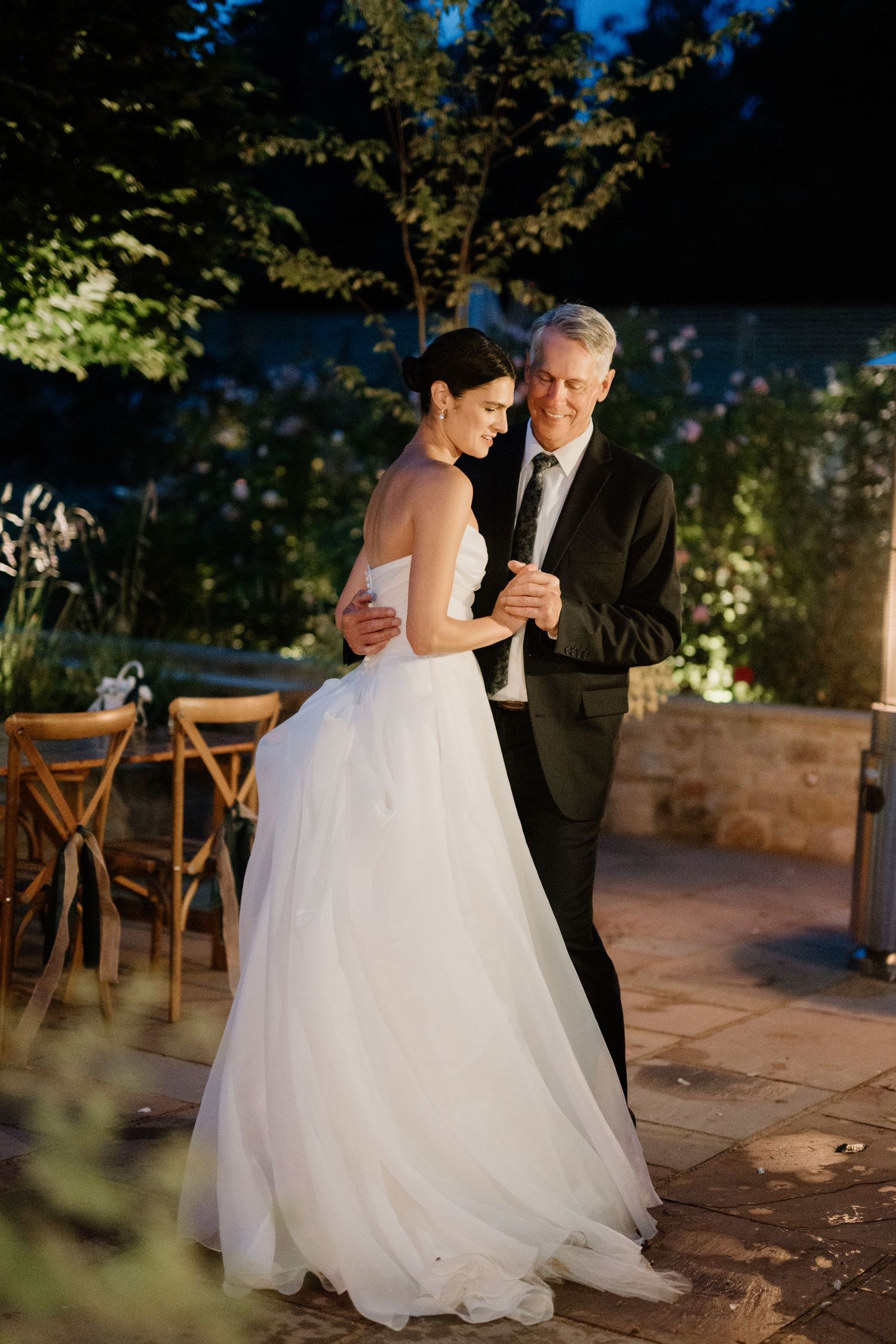 Bride dancing with older gentleman in black suit at outdoor evening reception.