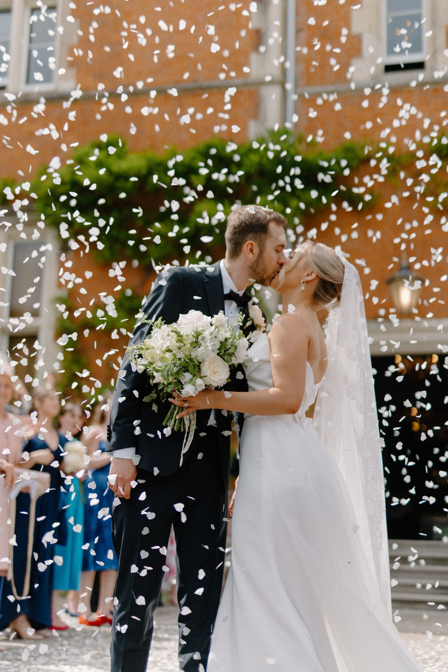 Newlyweds share a kiss while white confetti petals fall around them outside a brick building.