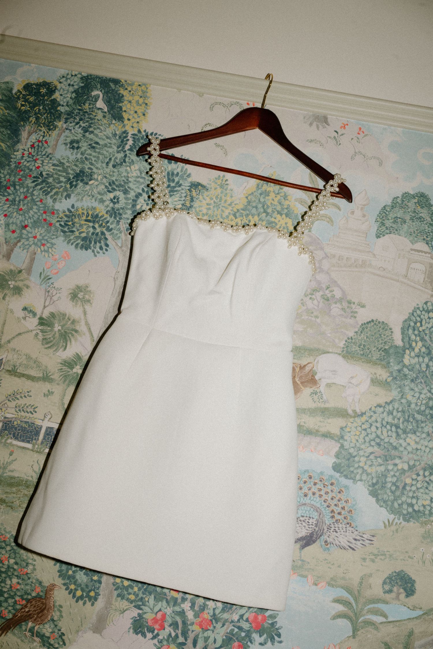 Elegant white mini wedding dress with pearl trim straps hanging on a decorative hanger.