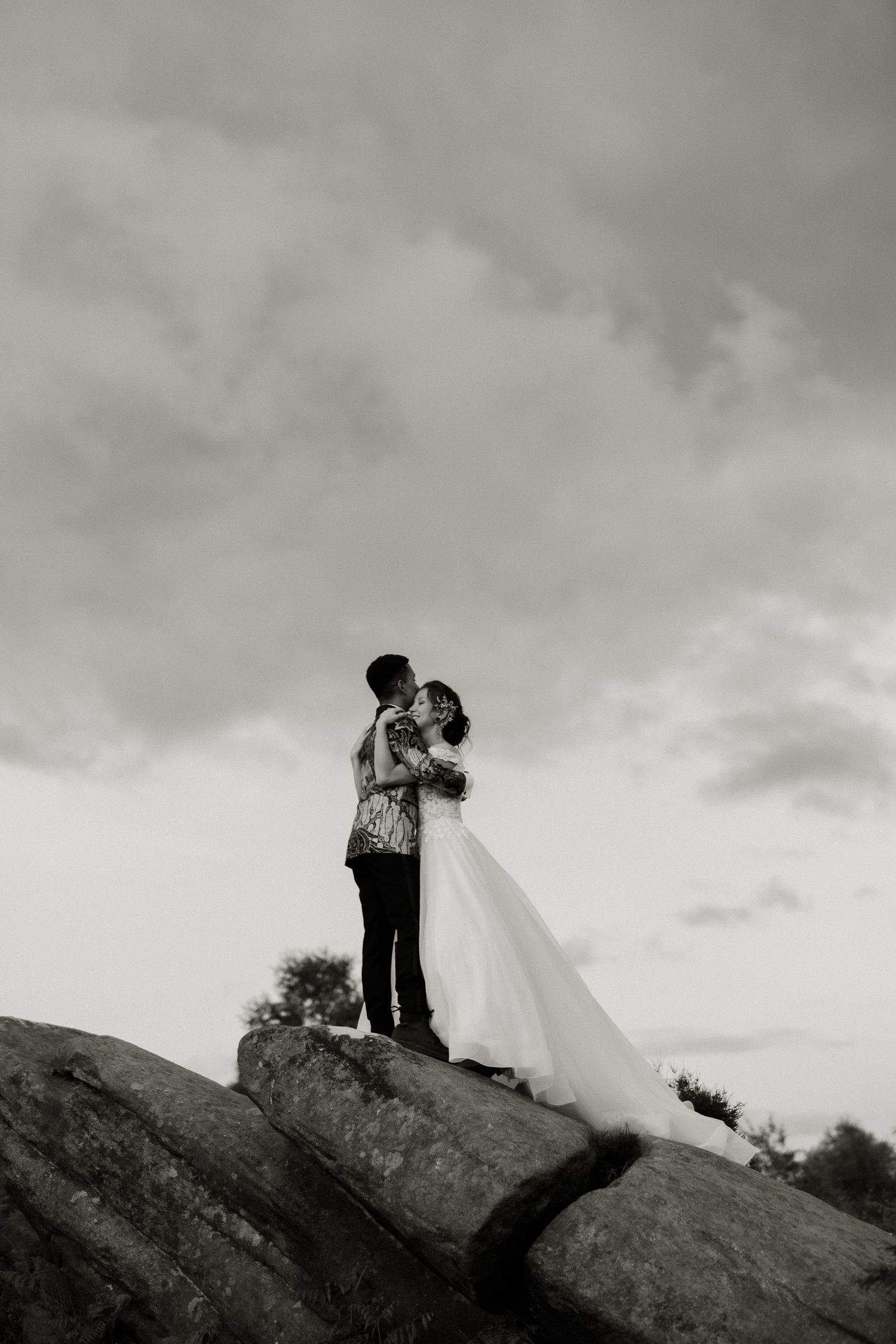 Black and white photo of bride and groom kissing on rocky outcrop beneath dramatic cloudy sky.