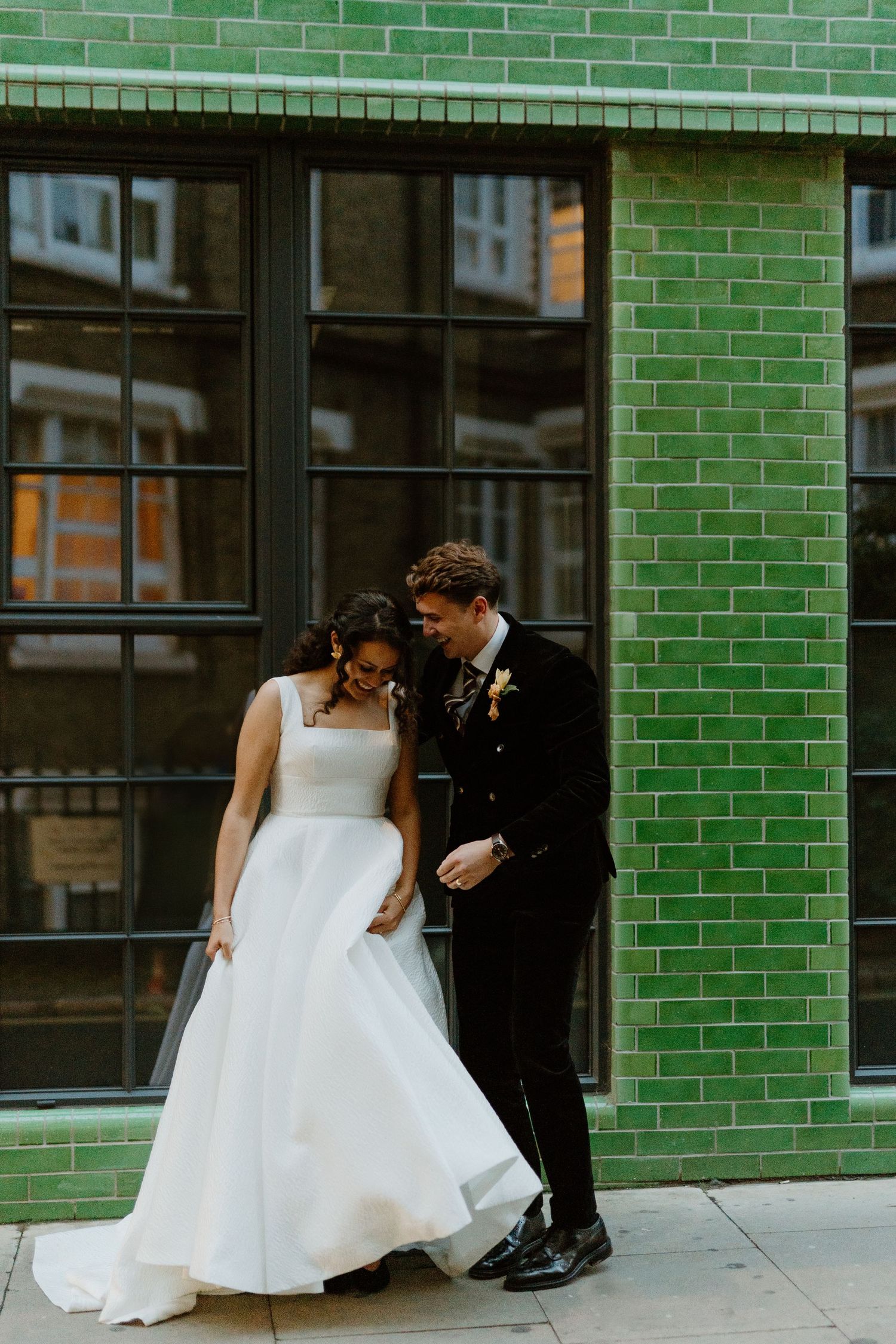 Bride and groom laughing together outside green tiled building on their wedding day in London.