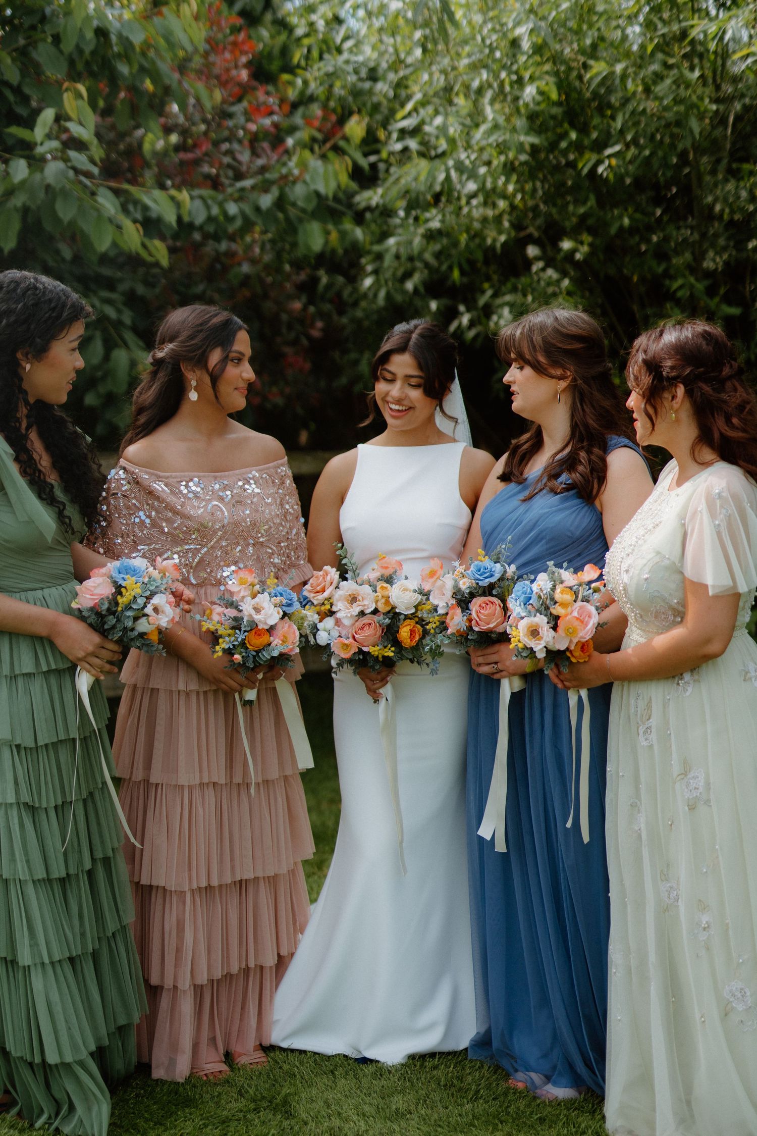 Bride and bridesmaids in pastel dresses laughing together while holding colorful floral bouquets.